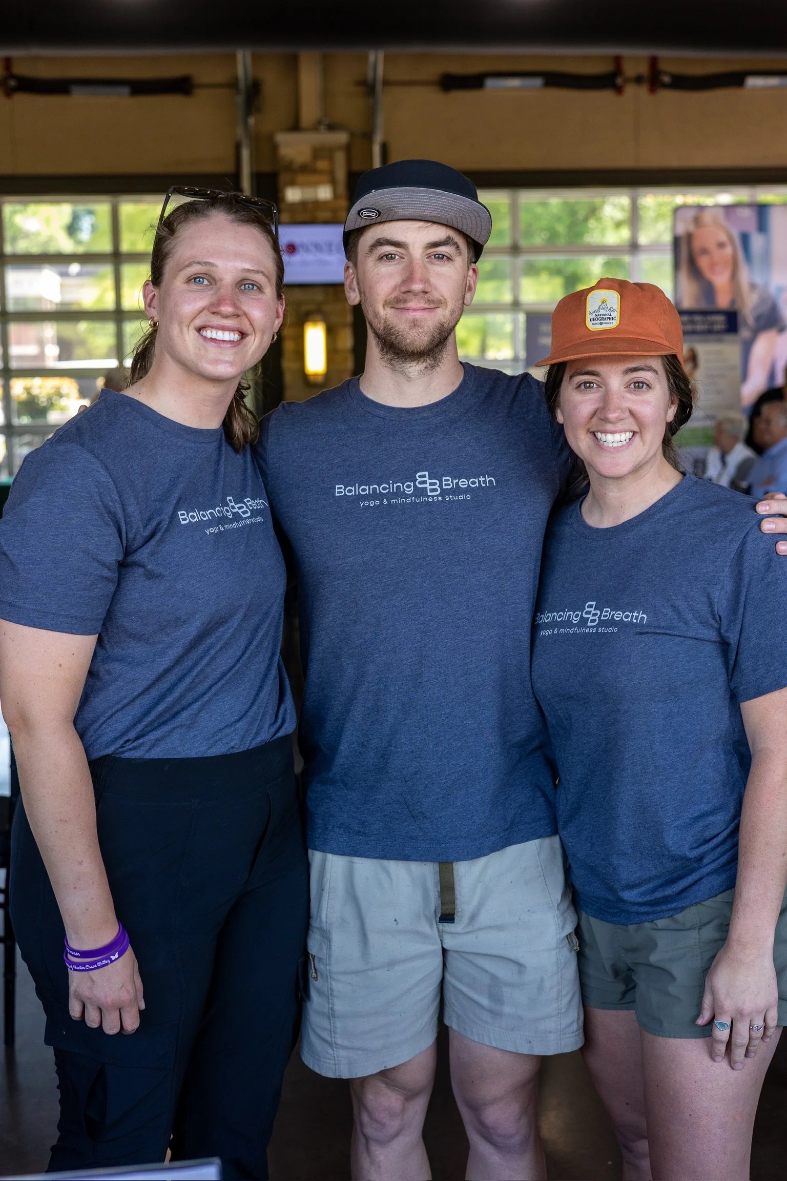 Three people wearing matching blue T-shirts with 'Balancing Breath' logo, standing indoors with bright windows, smiling for the camera.