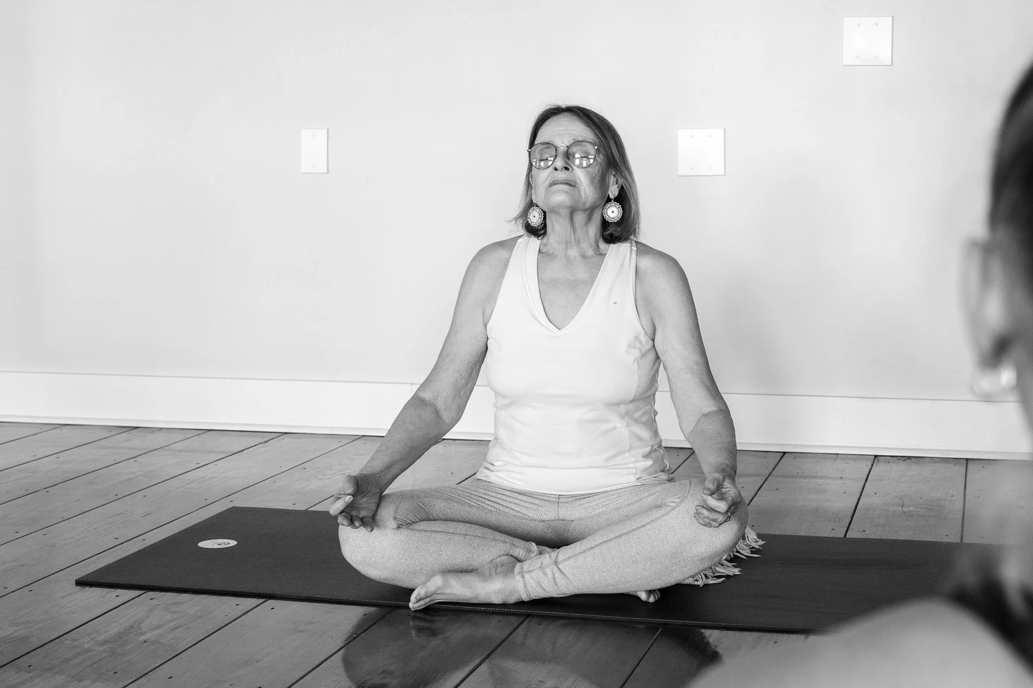 An elderly woman practicing yoga indoors, seated in a cross-legged meditation pose on a mat, with her hands resting on her knees and fingers forming a mudra, wearing glasses, earrings, and a tank top.