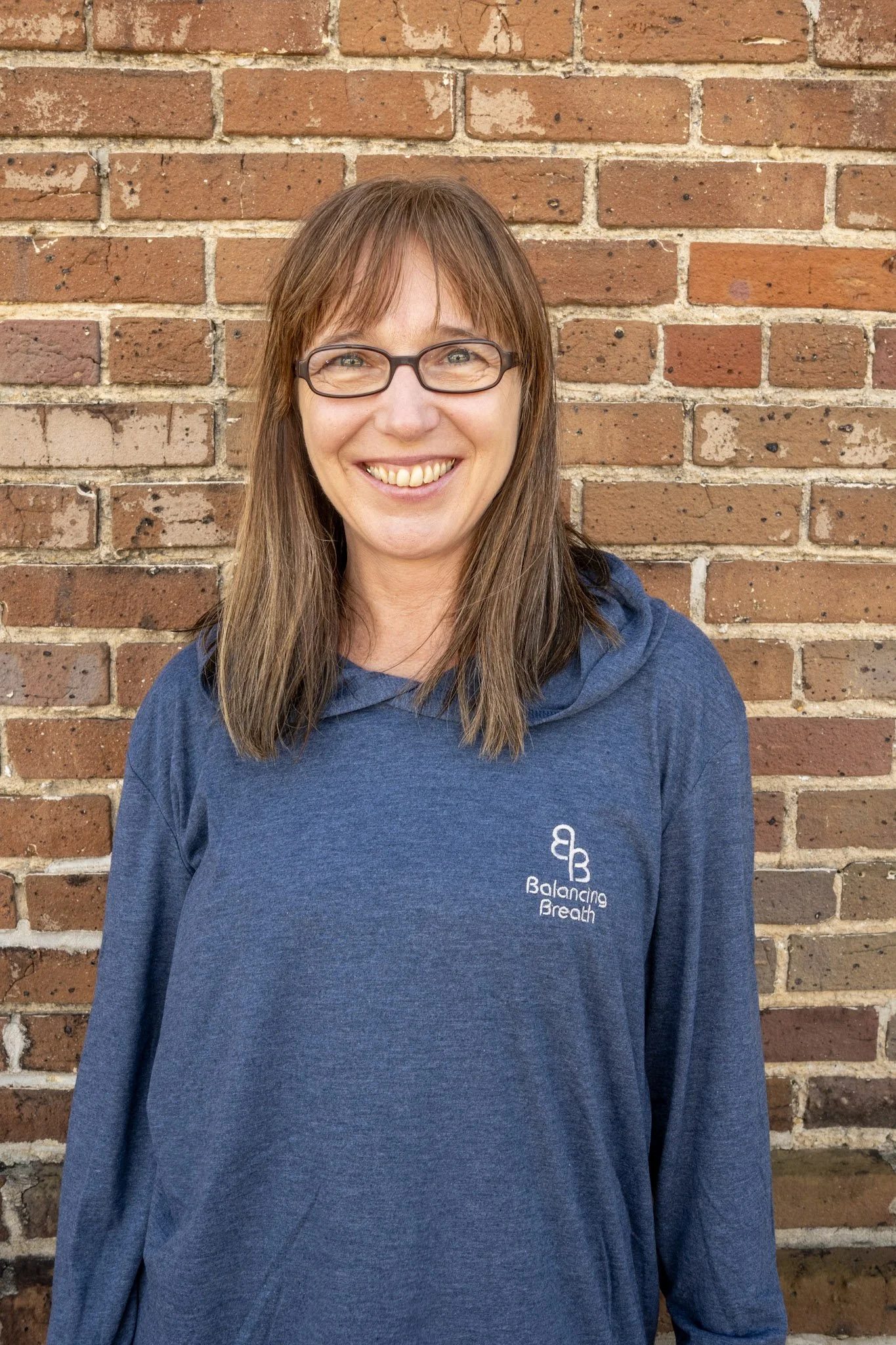 A woman with glasses and brown hair smiling while holding colorful exercise balls against a brick wall background.