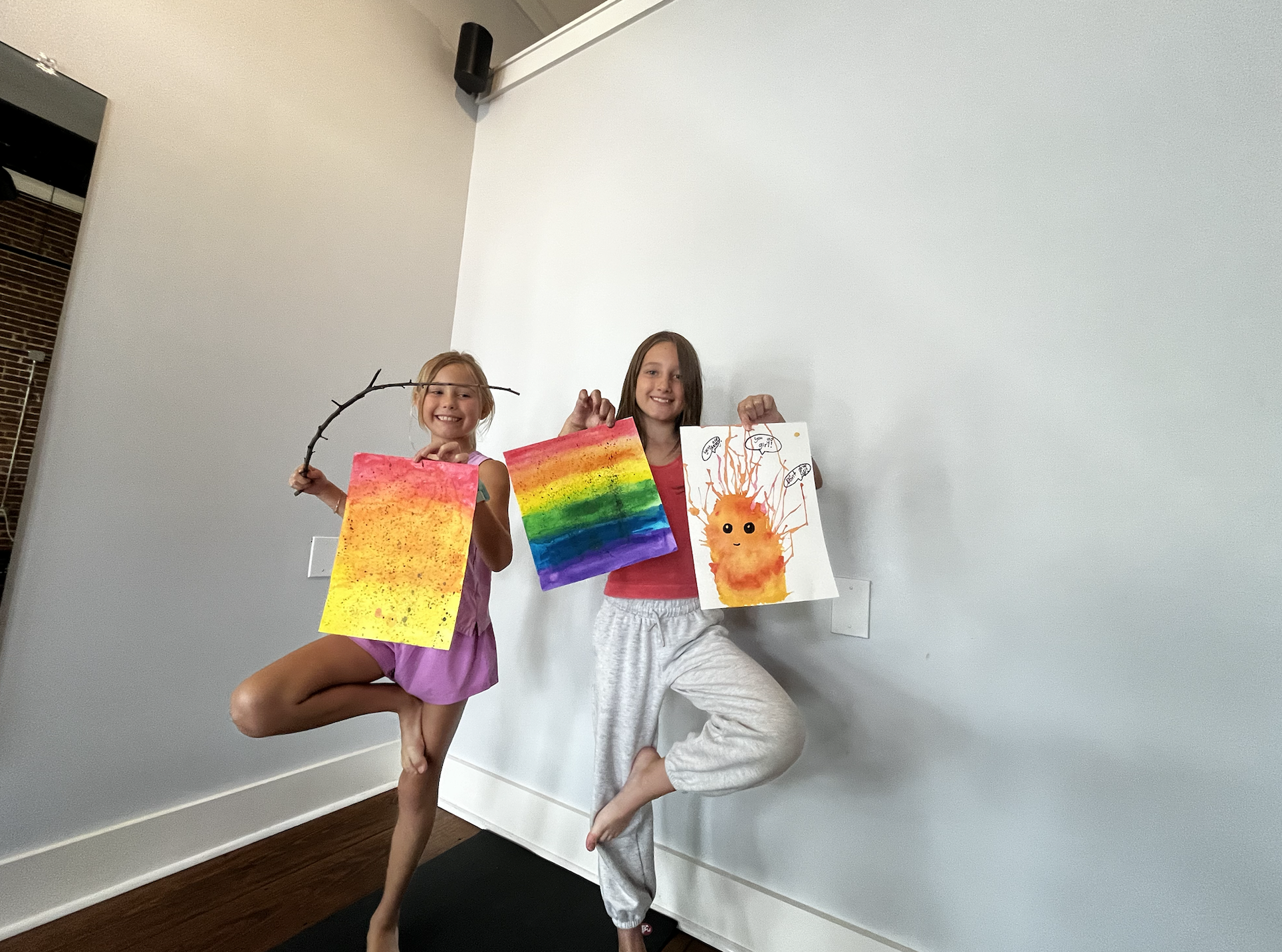 Two young girls happily display colorful artwork: one holding a rainbow painting and the other holding a picture of a fluffy orange creature with colorful streamers, in a bright room.