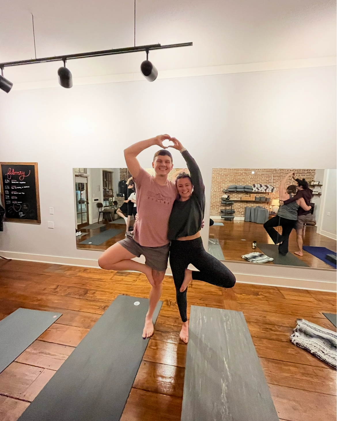 Two people practicing yoga and balancing on one leg each, forming a heart shape with their arms, in a yoga studio with hardwood floors and yoga mats.