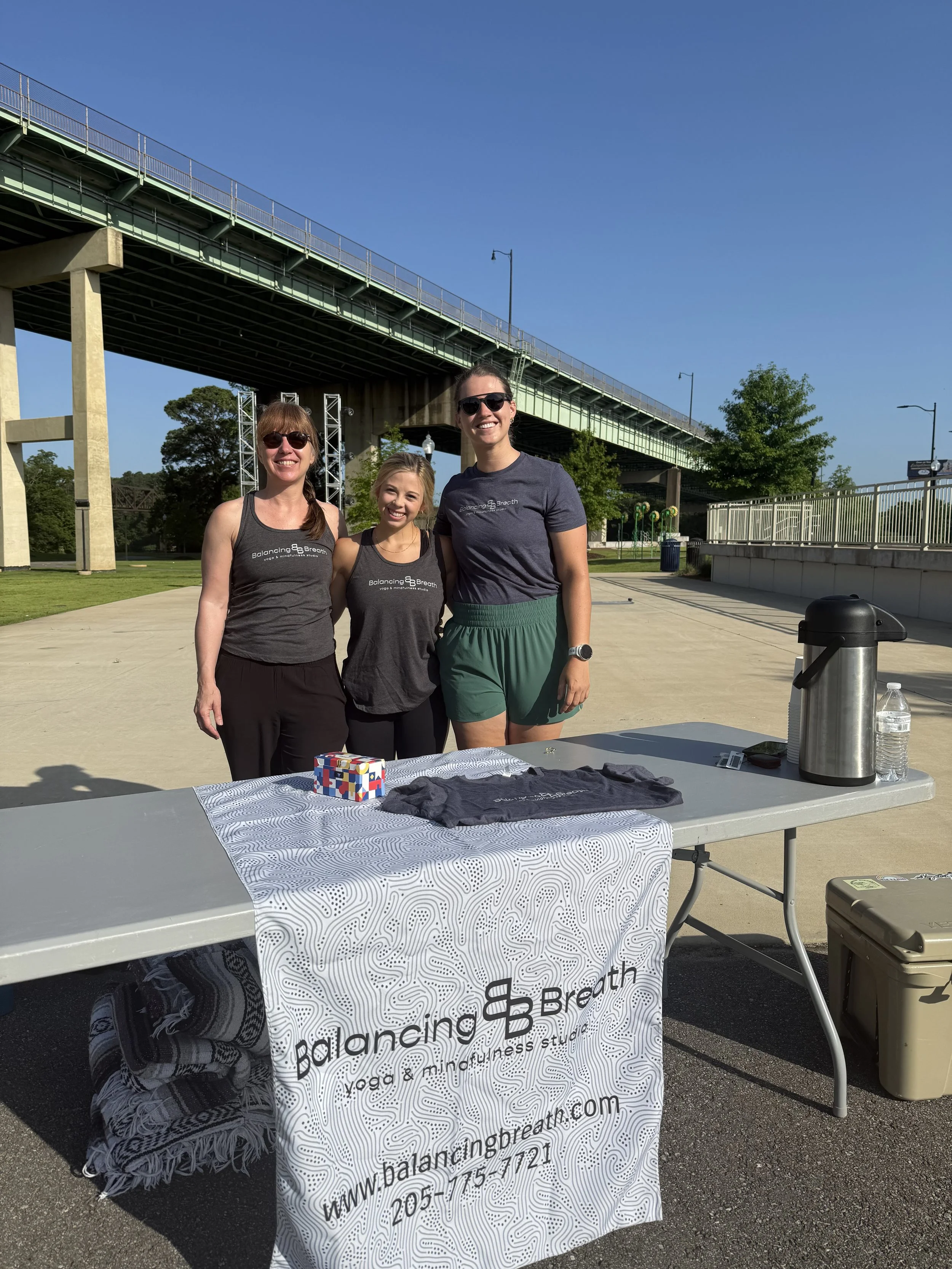 Three women standing behind a table at an outdoor event, under a bridge with blue sky and green trees in the background. The table has a banner for Balancing Breath yoga and mindfulness studio.