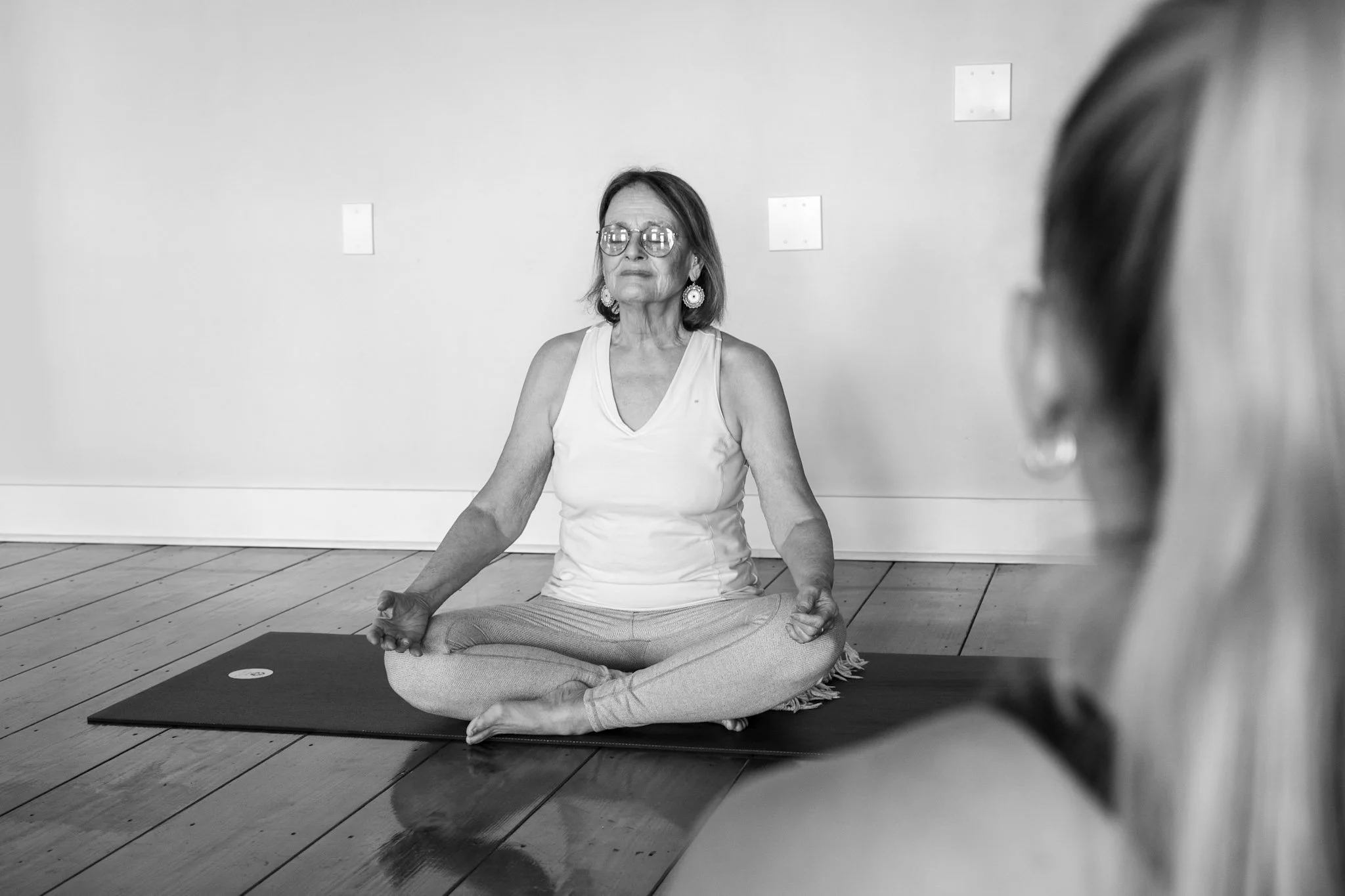 A woman practicing yoga or meditation on a mat while sitting cross-legged in a room with wooden floor and plain wall.