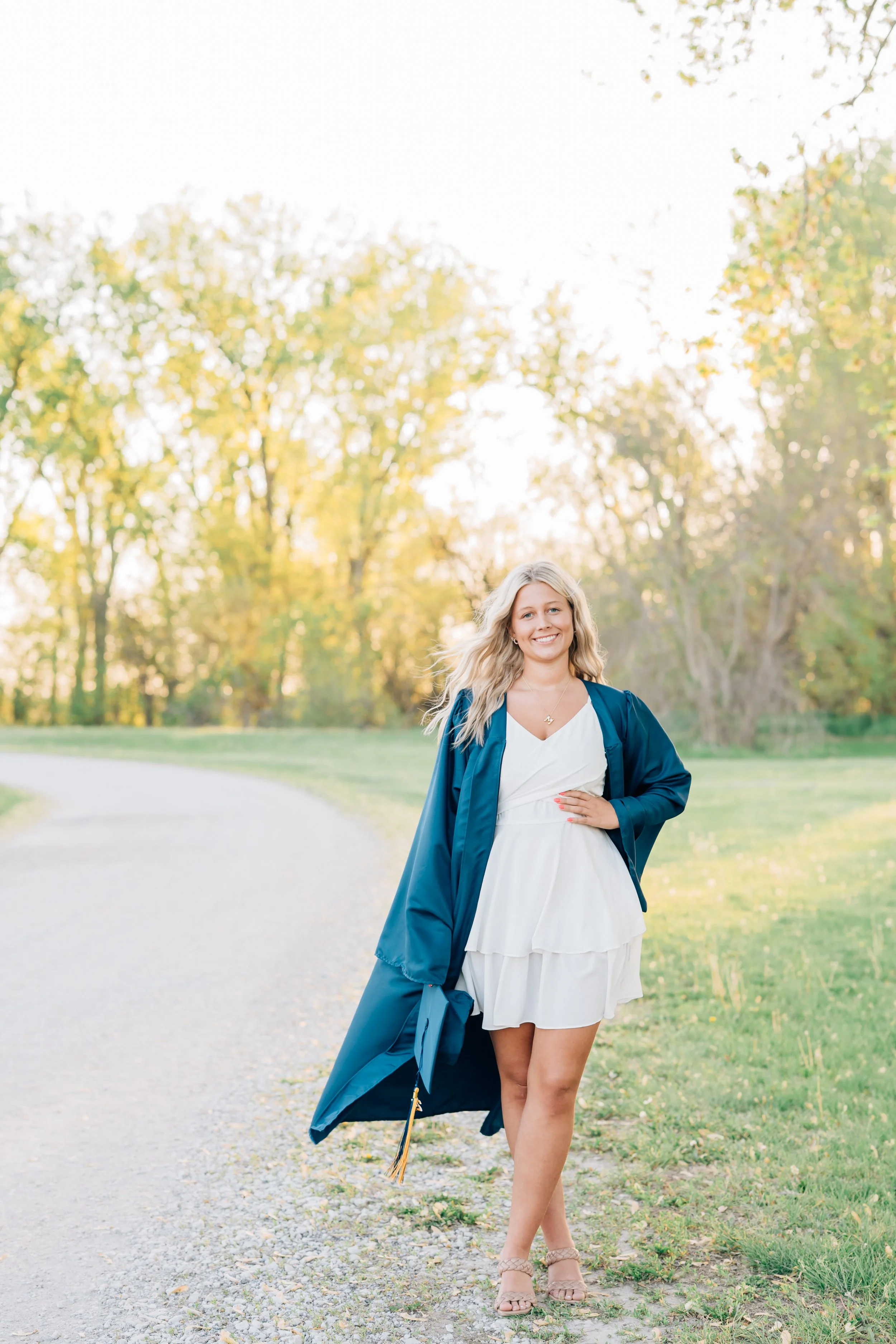 Girl poses in blue cap and gown with white dress celebrating her graduation during cap and gown photos. 