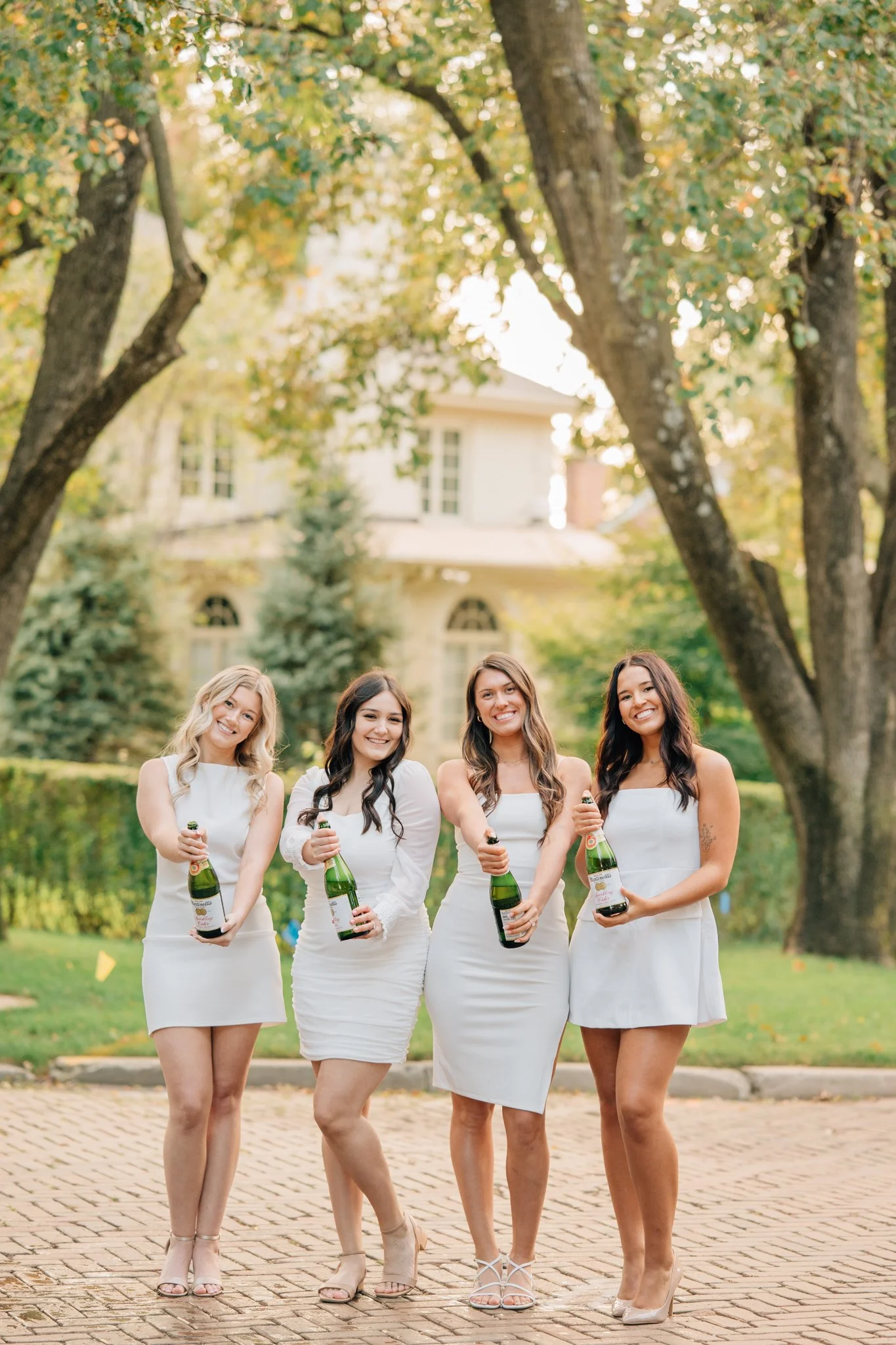 Four nursing graduates in white dresses pose with champagne bottles before they pop them to celebrate graduation during grad photo session in Omaha.