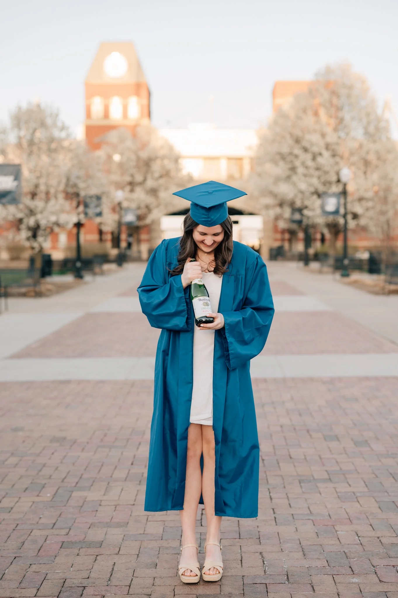 College graduation picture of girl in blue cap and gown posing on Creighton's campus, holding bottle of champagne.