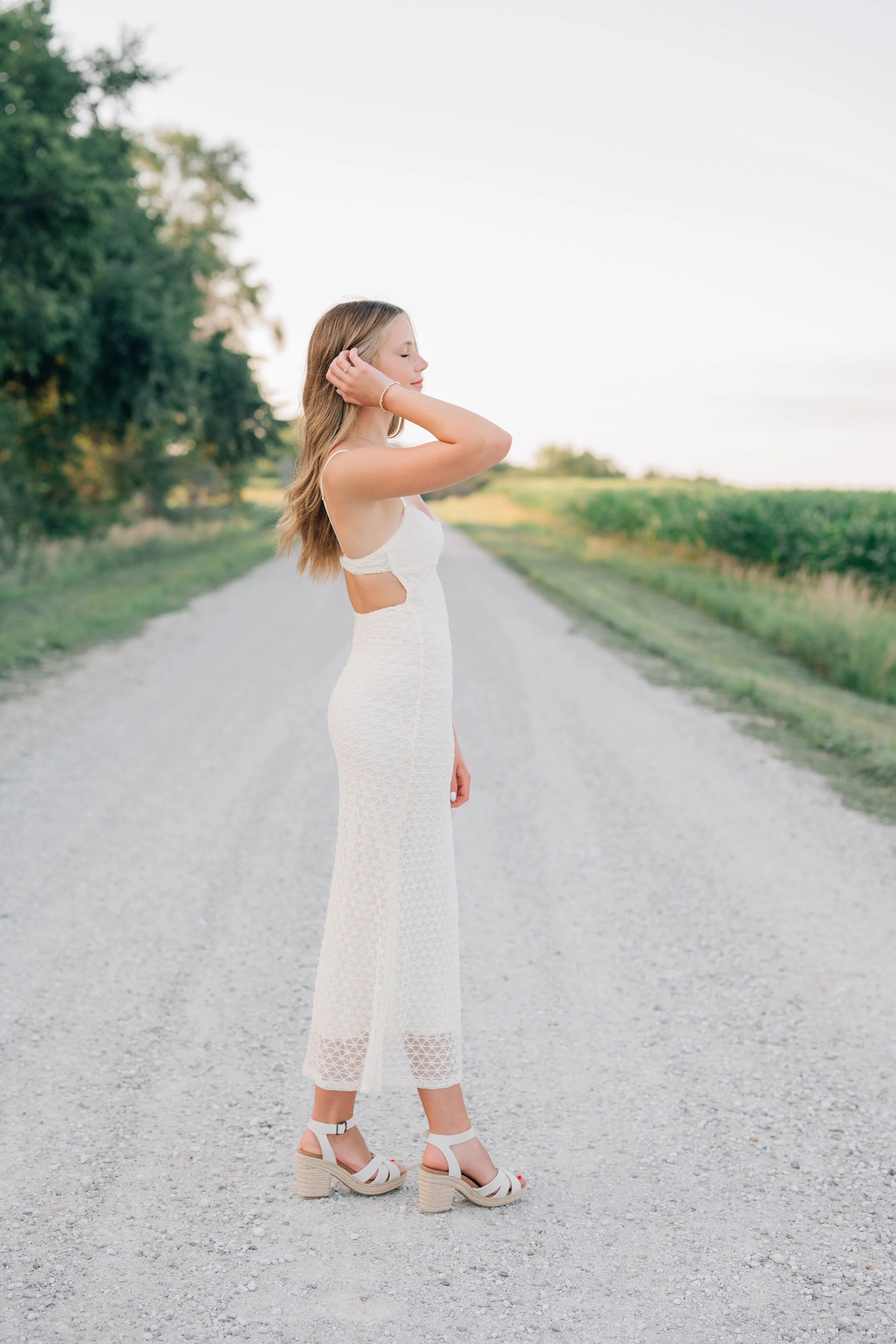 Candid photo of girl tucking her hair behind her ear, wearing white dress during senior photography session.