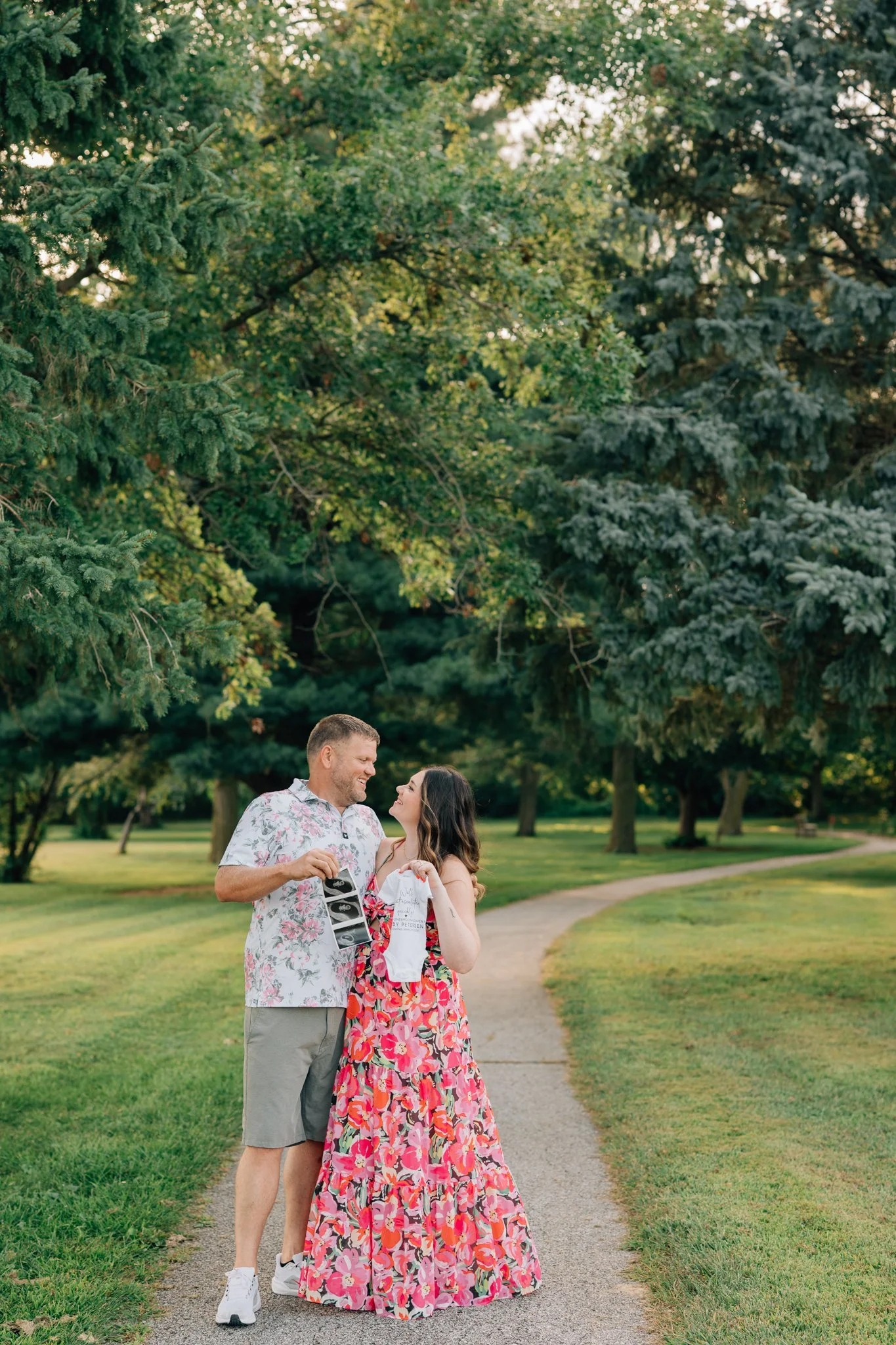 Couple cuddles in a park holding a baby onesie to announce pregnancy. 