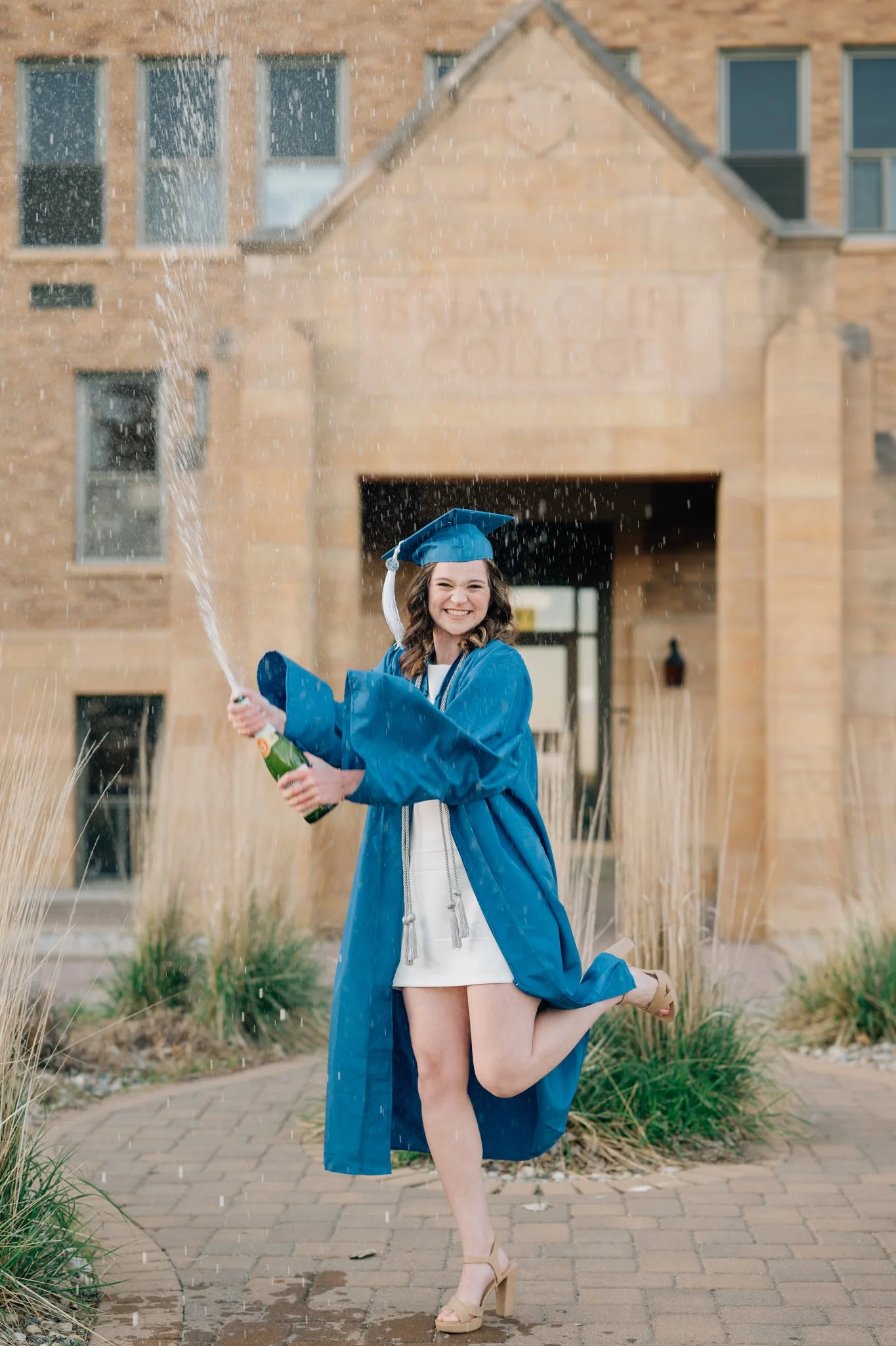 Girl in blue cap and gown pops bottle of champagne on campus during her cap & gown college graduation photo session.