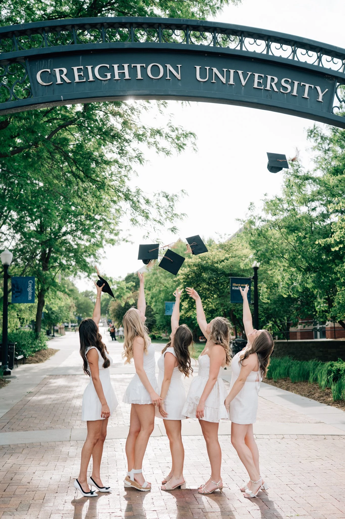 5 friends toss caps into the air under the Creighton University arch on campus in Omaha, NE during their graduation picture session.