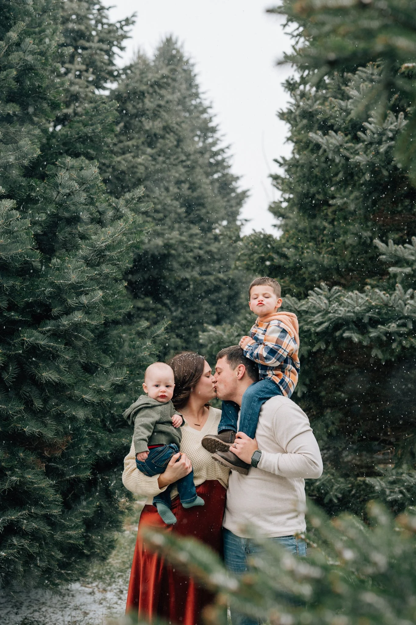 Couple cuddles among pine trees with their boys looking to camera during winter family photography session. 