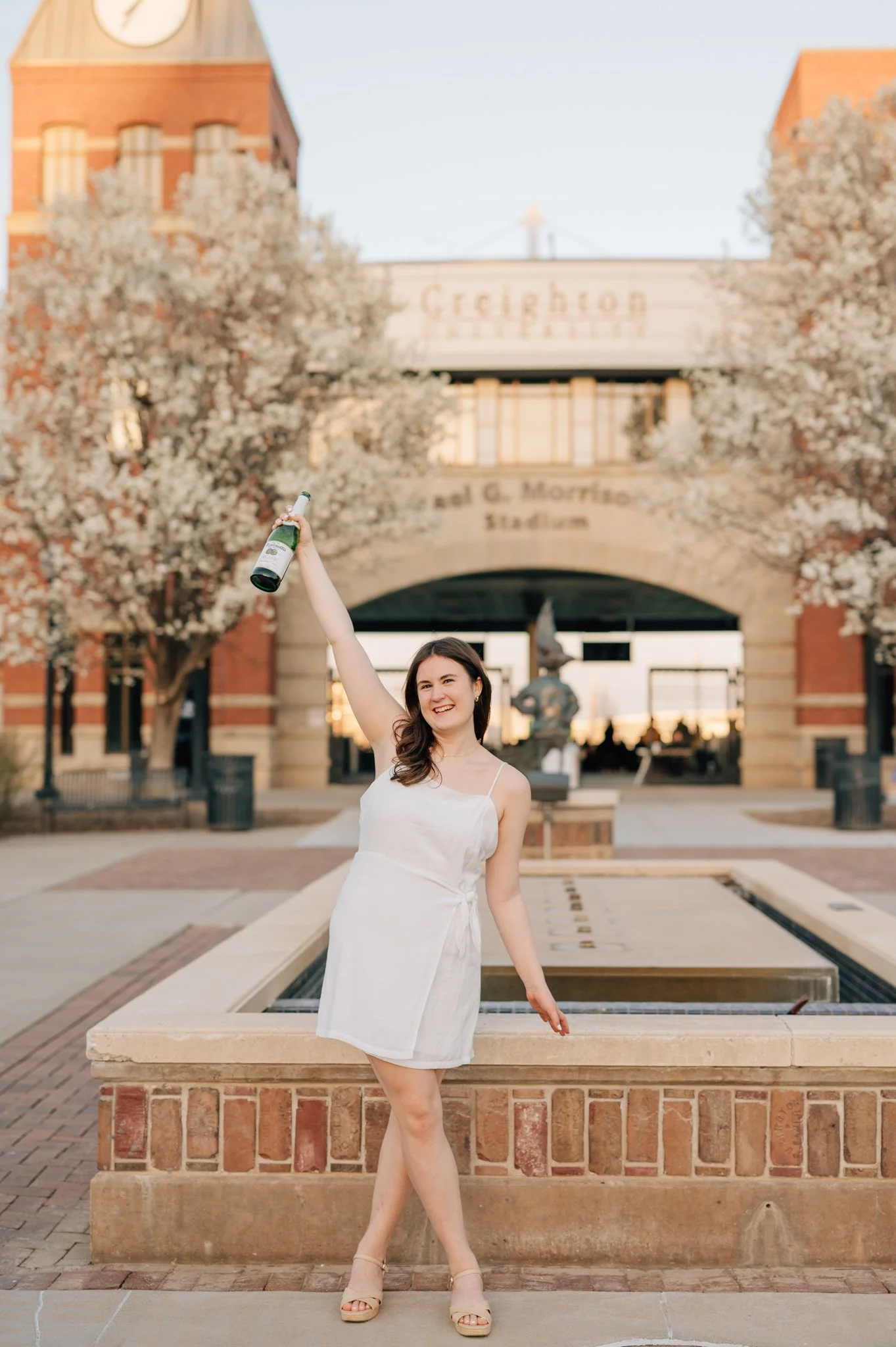 College graduate in white dress raises bottle of champagne to celebrate graduation from Creighton University during cap and gown photos.