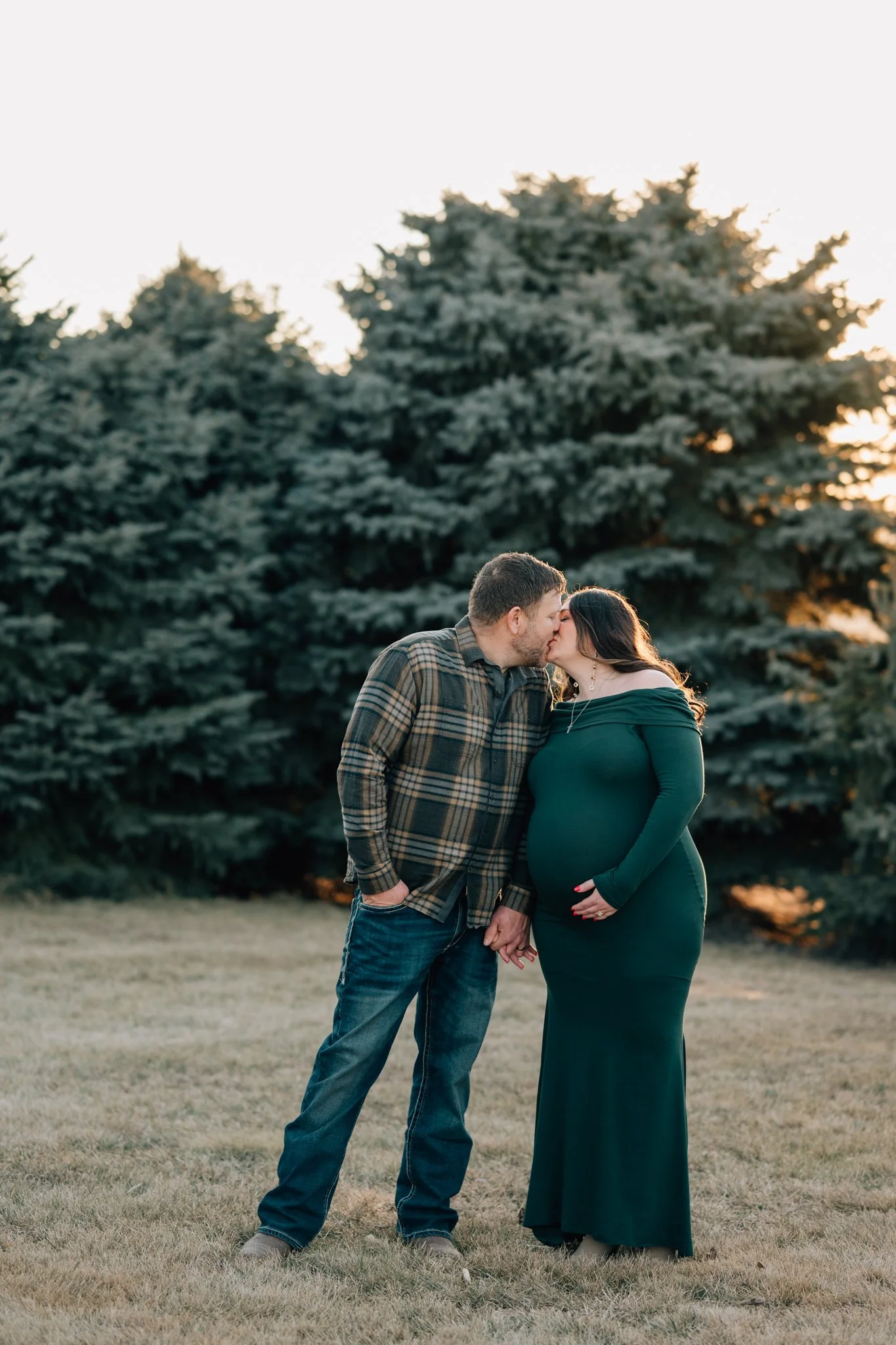 Couple poses for maternity photos in Iowa during winter, surrounded by pine trees.
