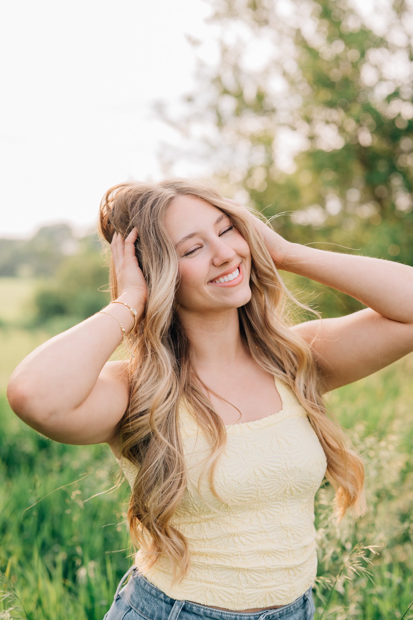 Senior girl smiles and plays with hair in a grassy field for senior pictures.
