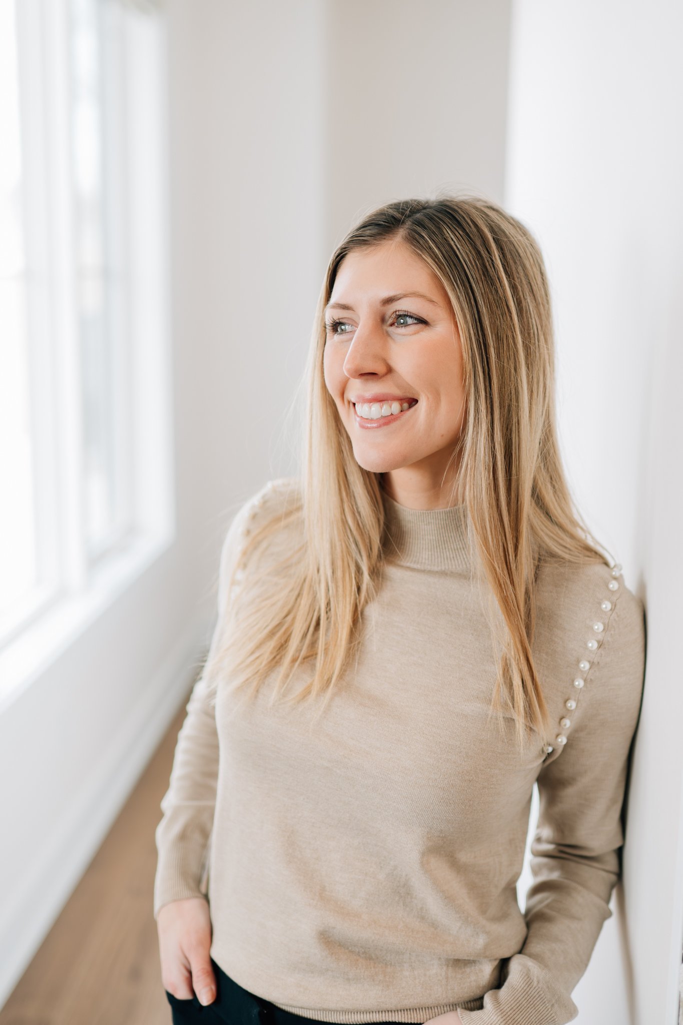Professional headshot of a realtor, leaning against a white wall, looking out windows for her branding photography session.