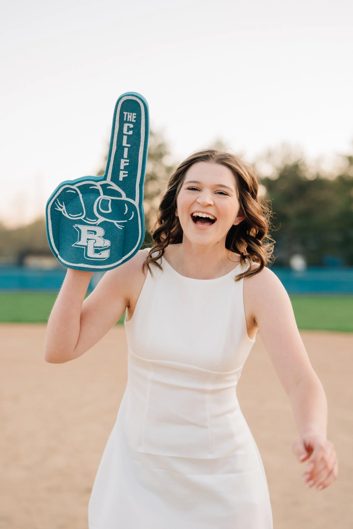 Girl laughs to camera wearing a white dress and a foam finger to celebrate her college graduation pictures.
