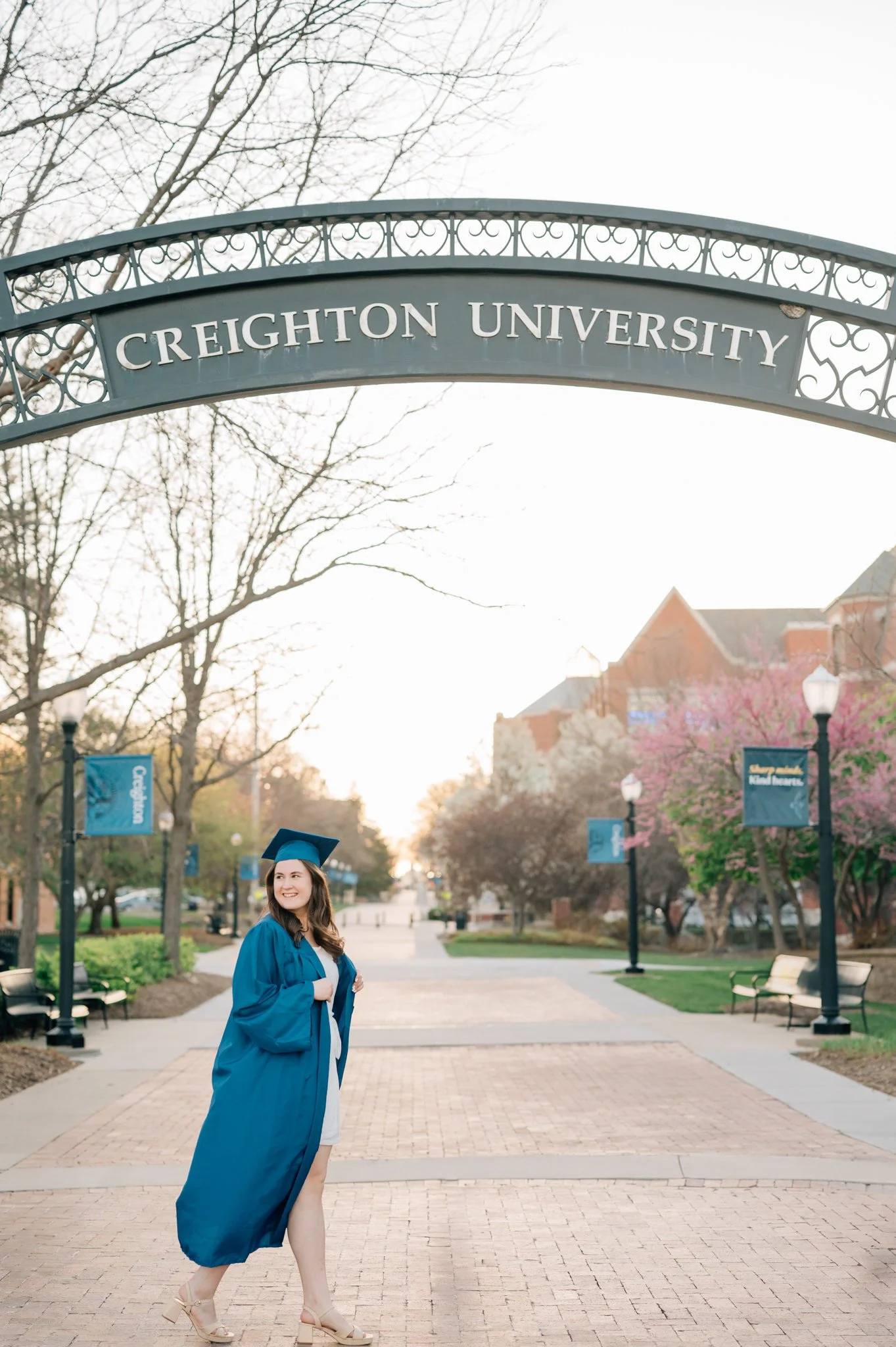 Girl in blue cap and gown walks under Creighton University sign during her college graduation photography session.