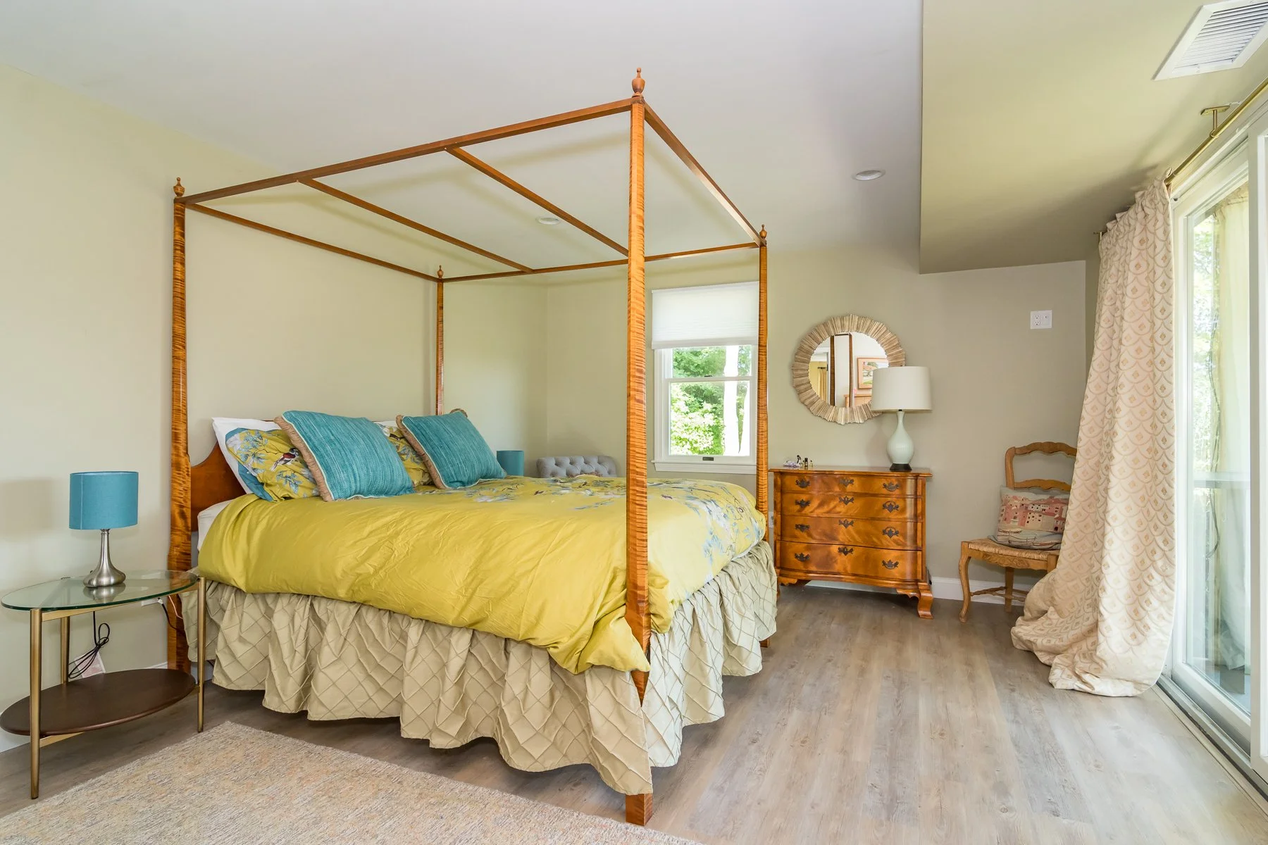 Bedroom with a wooden four-poster bed, yellow bedding, blue pillows, a glass nightstand with a blue lamp, a wooden chest of drawers with a white lamp, a chair, mirror, curtain, and window with greenery outside.