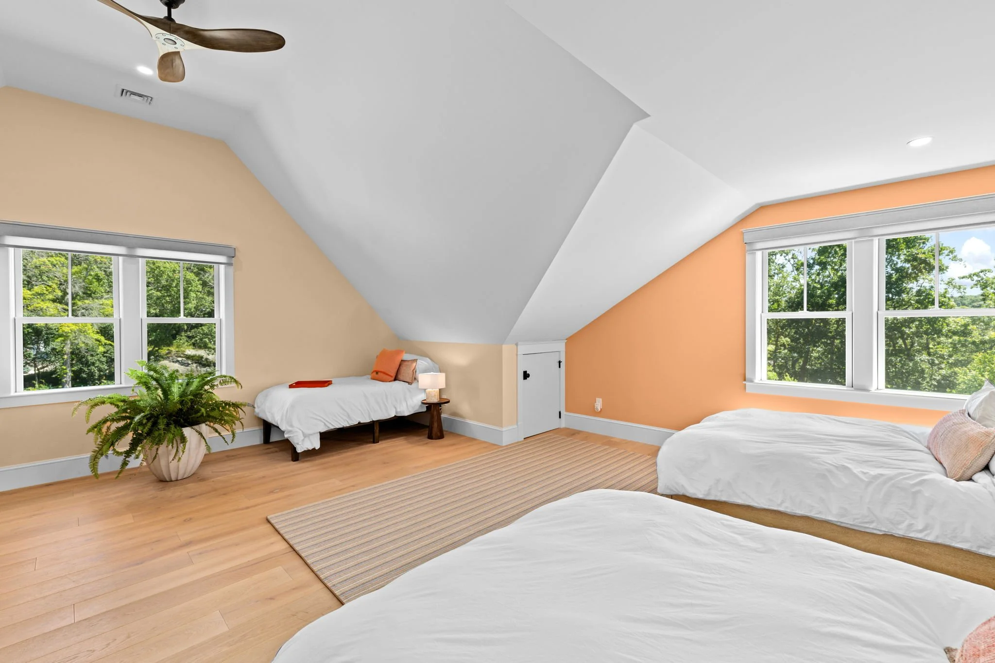 A bright bedroom with three beds with white linens, two large windows with green trees outside, a beige and orange accent wall, hardwood floors, a small ledge with a closet door, and a potted plant.