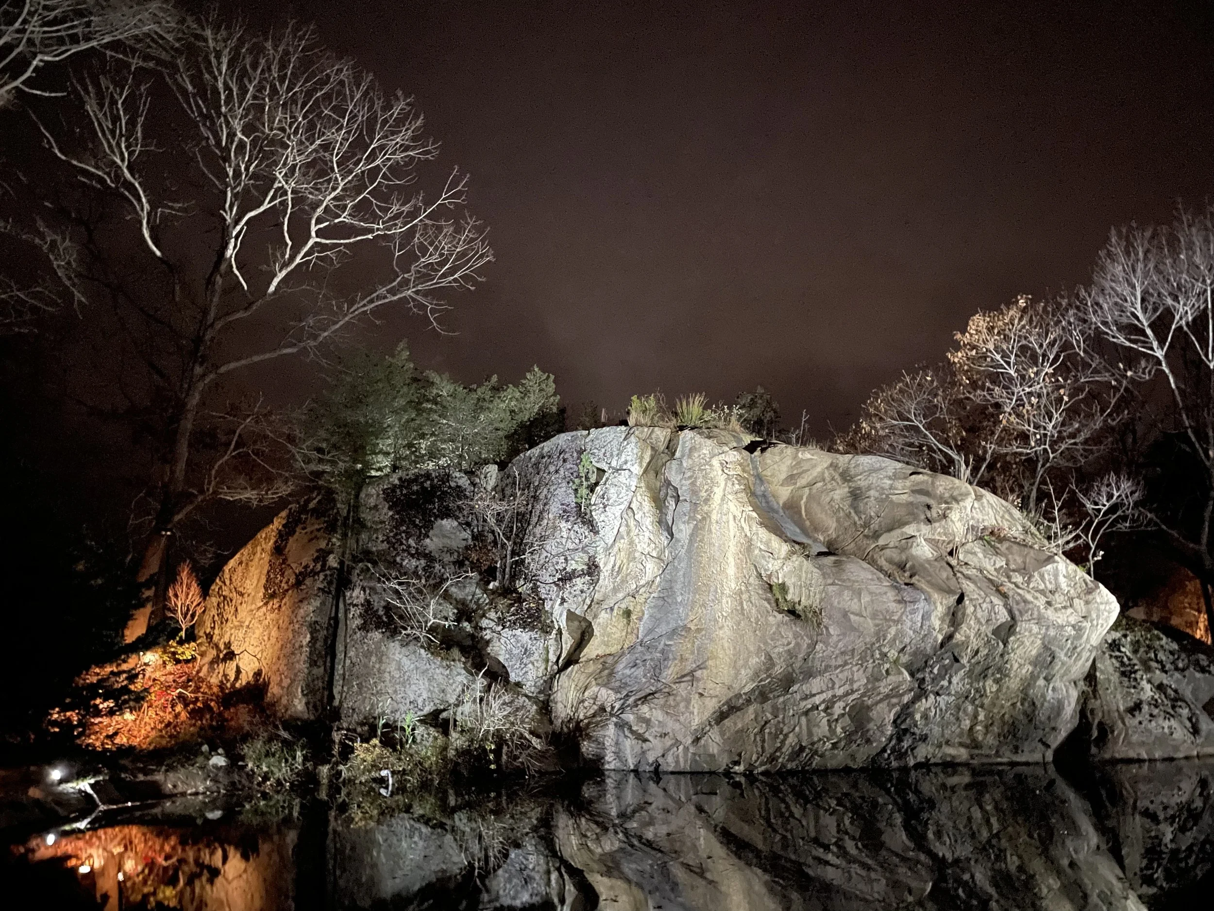 Nighttime scene of a large rock formation with trees around it, reflected in a body of water below, illuminated by artificial lighting.