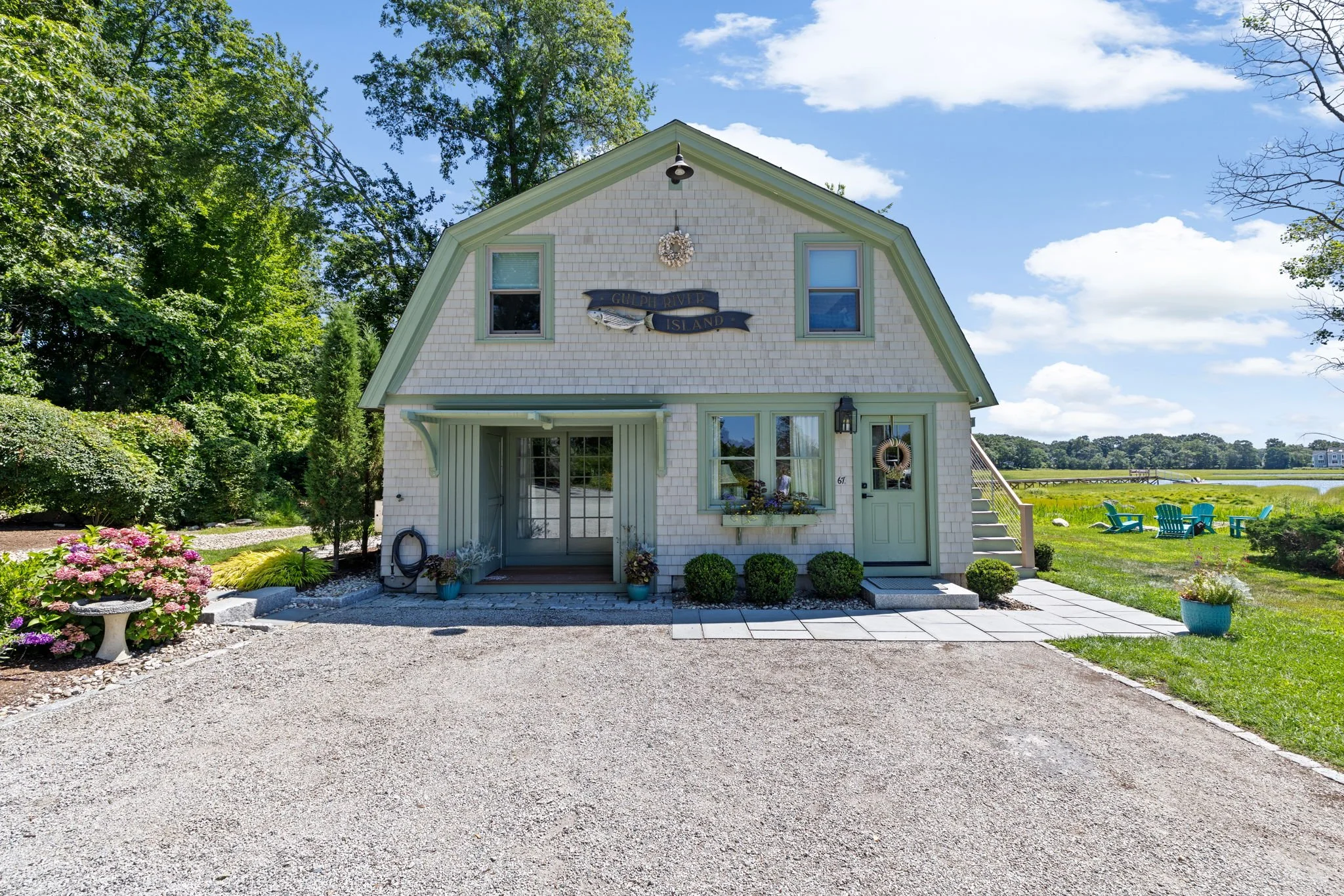 A charming two-story house with white shingle siding, surrounded by lush greenery and a gravel driveway. The house has a small front porch, decorative shutters, and a sign that reads "Gulph River Island." There are potted plants, a wreath on the door