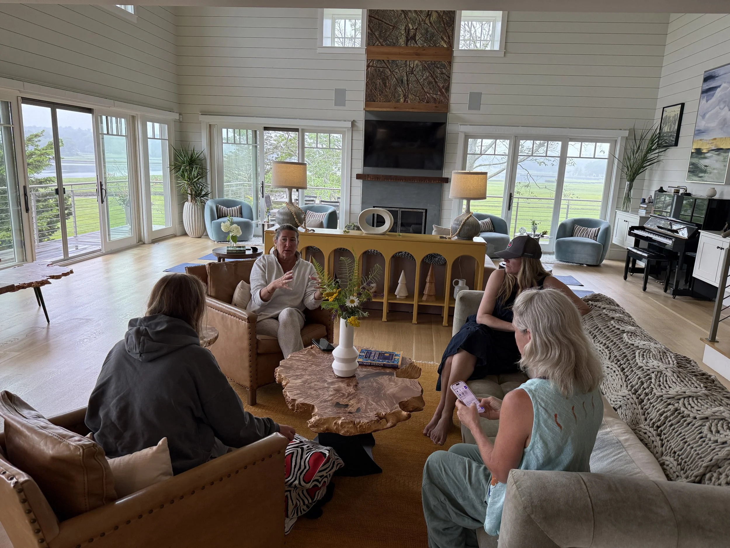 Group of five women having a discussion in a bright living room with large windows and outdoor view.