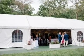 Large white event tent with arched windows, set up outdoors with people inside attending a gathering or celebration, surrounded by trees and grass.