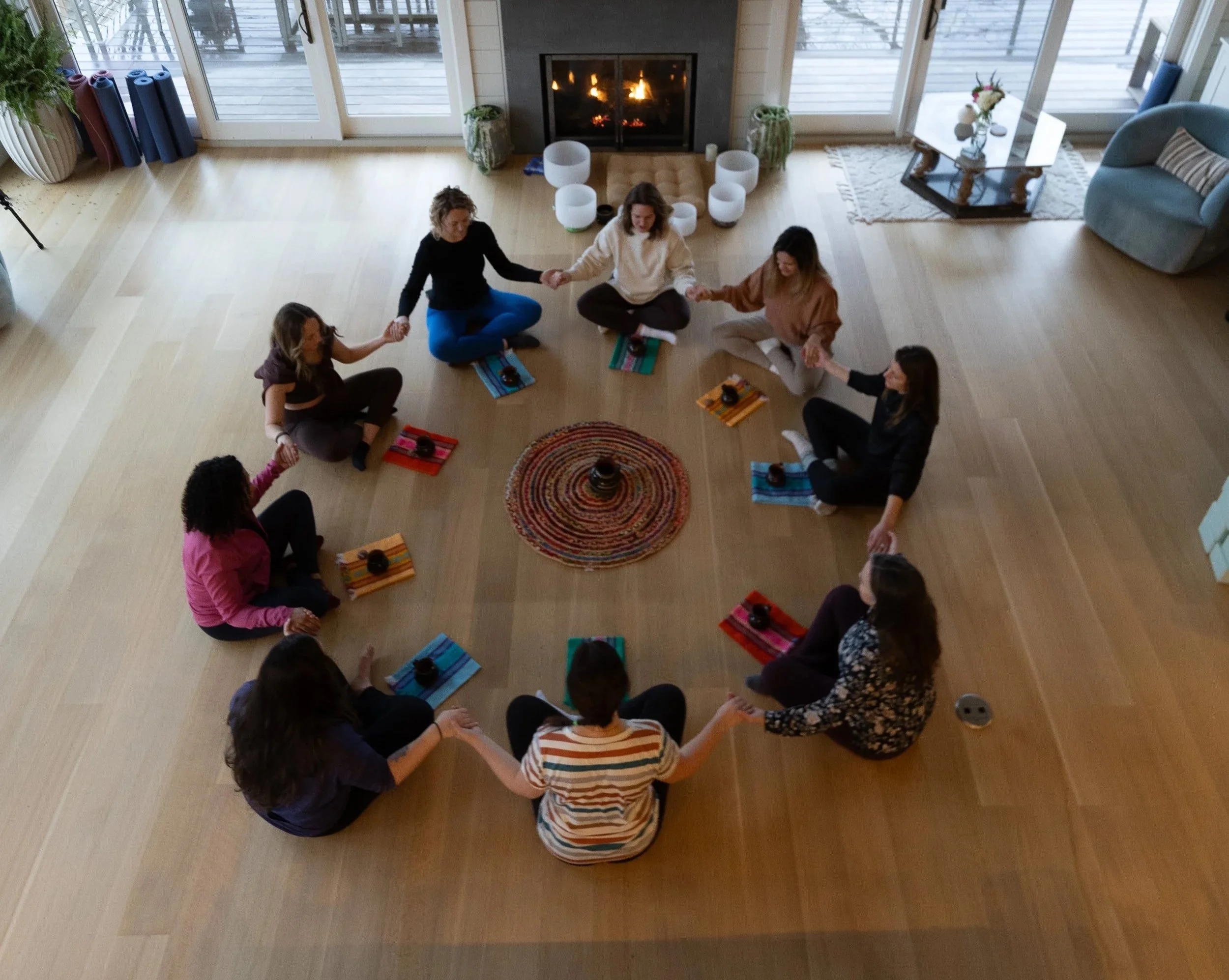 A group of nine women sitting in a circle holding hands in a cozy living room with a fireplace and large windows showing an outdoor deck.