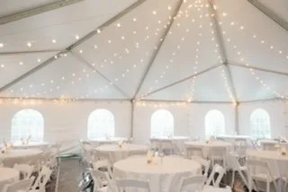 Inner view of a large event tent decorated with string lights, set up with round tables and chairs for a celebration.