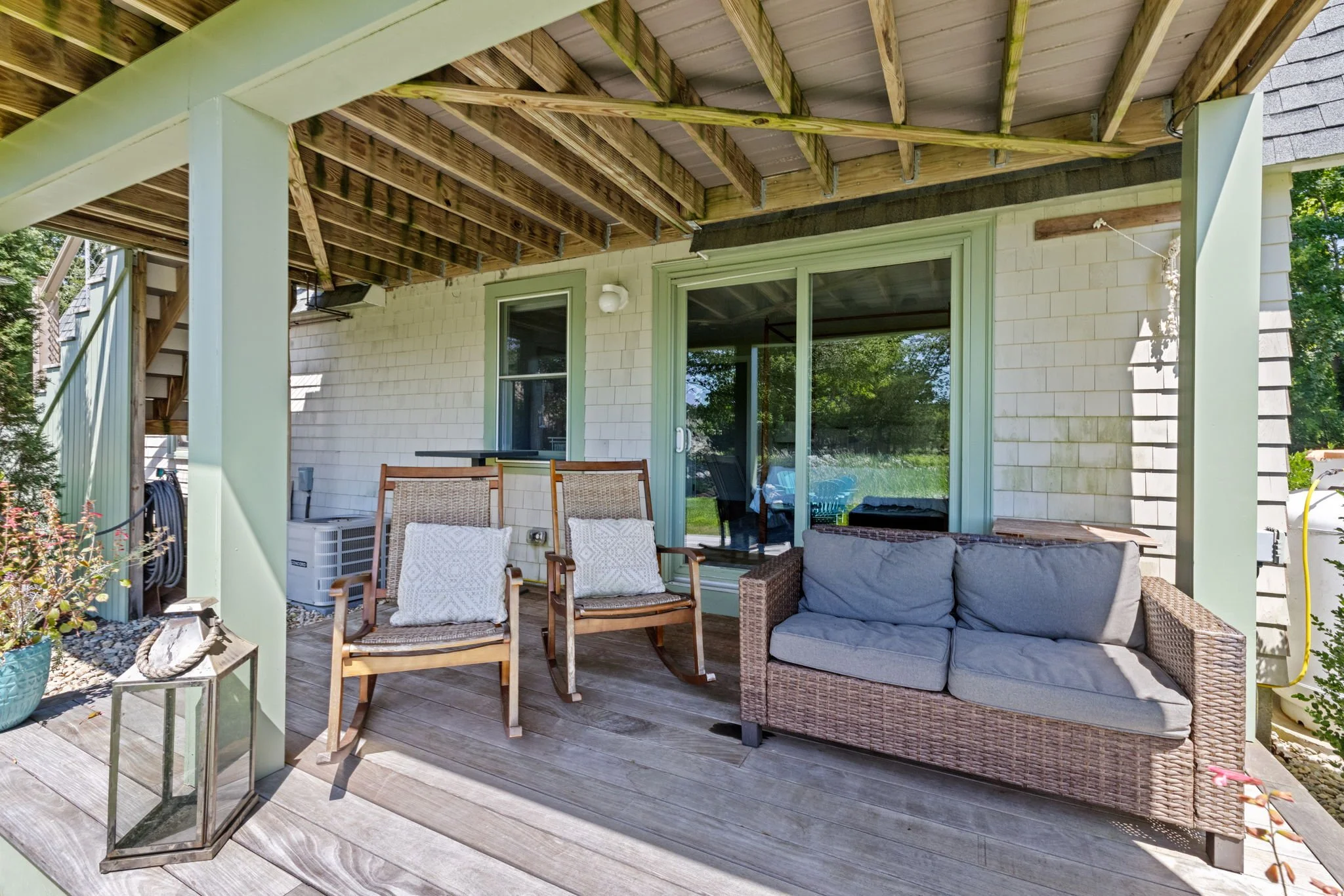 Outdoor patio with a three-piece wicker sofa, two wooden rocking chairs, and decorative lantern, surrounded by trees and shrubs.