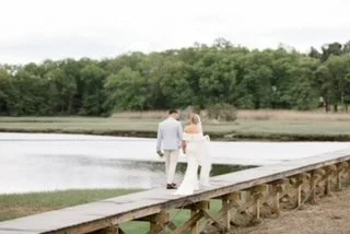 A couple walking hand in hand on a wooden dock near a body of water, with trees in the background.