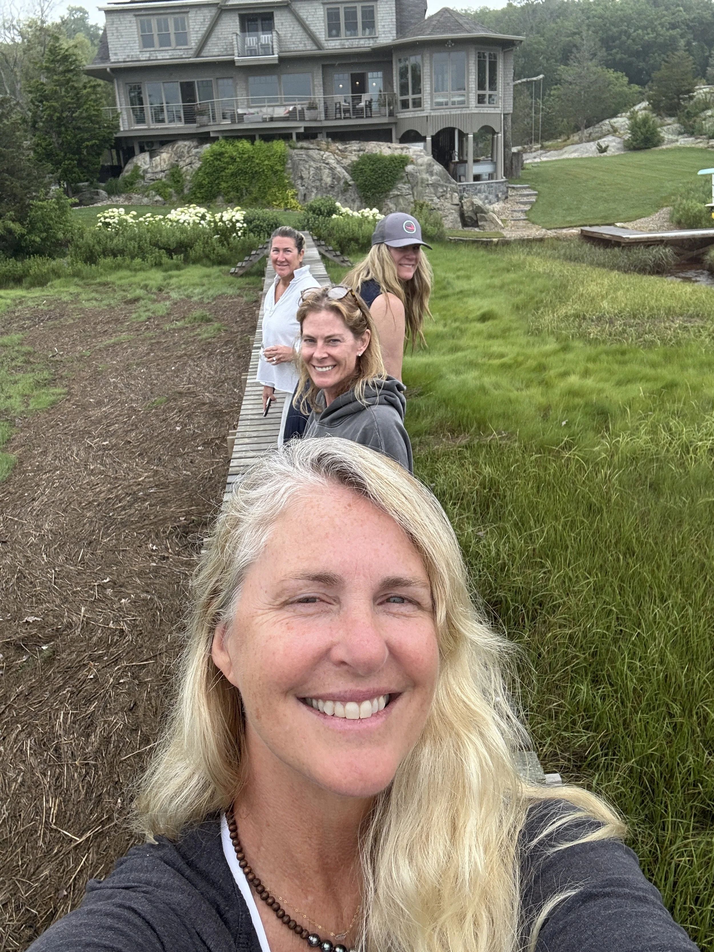 A group of six women stand on a narrow wooden dock, smiling and taking a selfie. They are outdoors near a large house with multiple levels and decks, surrounded by greenery and a well-maintained lawn. The woman in front has blonde hair and is smiling at the camera, with others lined behind her along the dock.