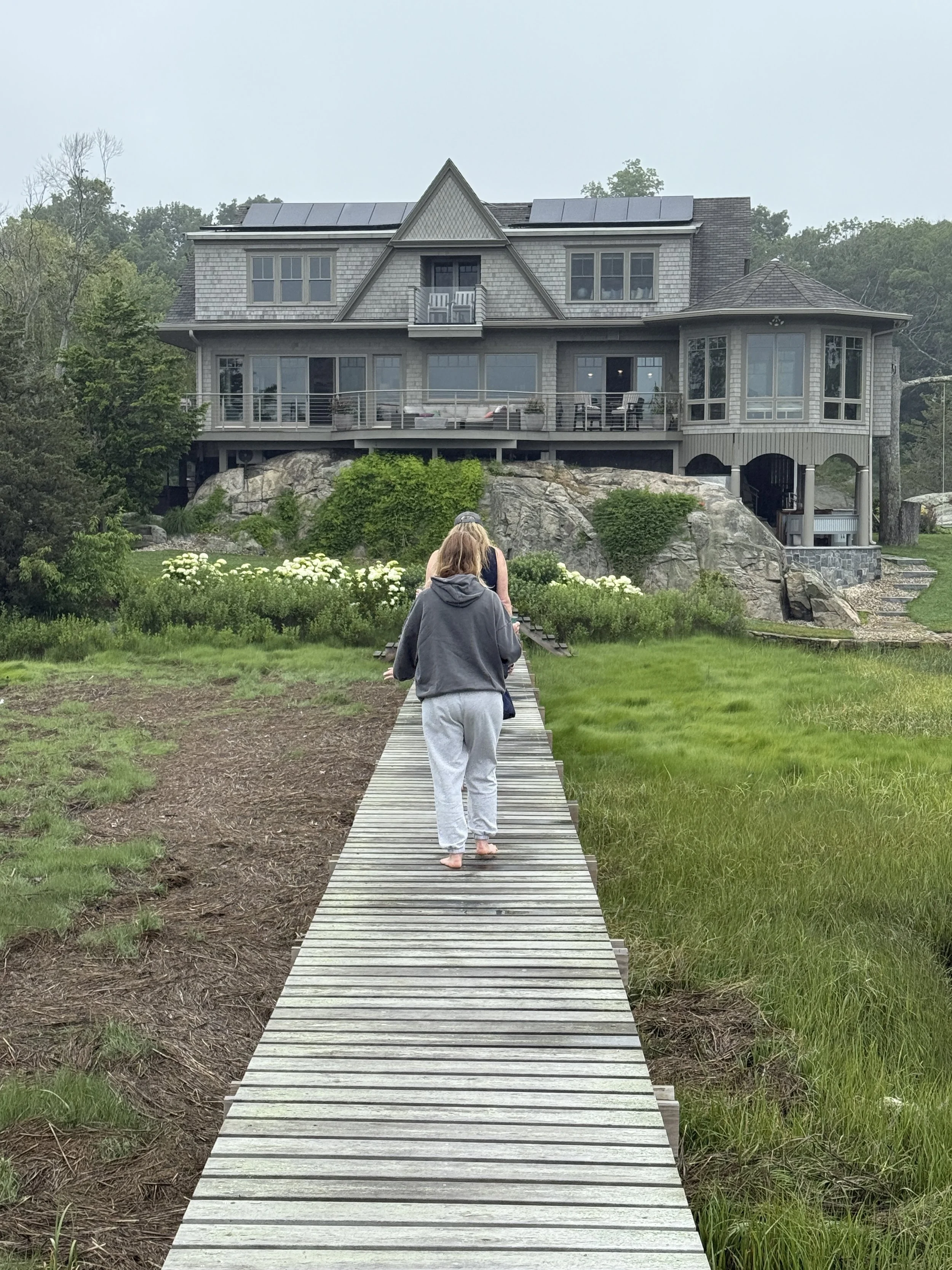 People walking on a wooden path towards a large modern house built on a rocky hill, surrounded by greenery.