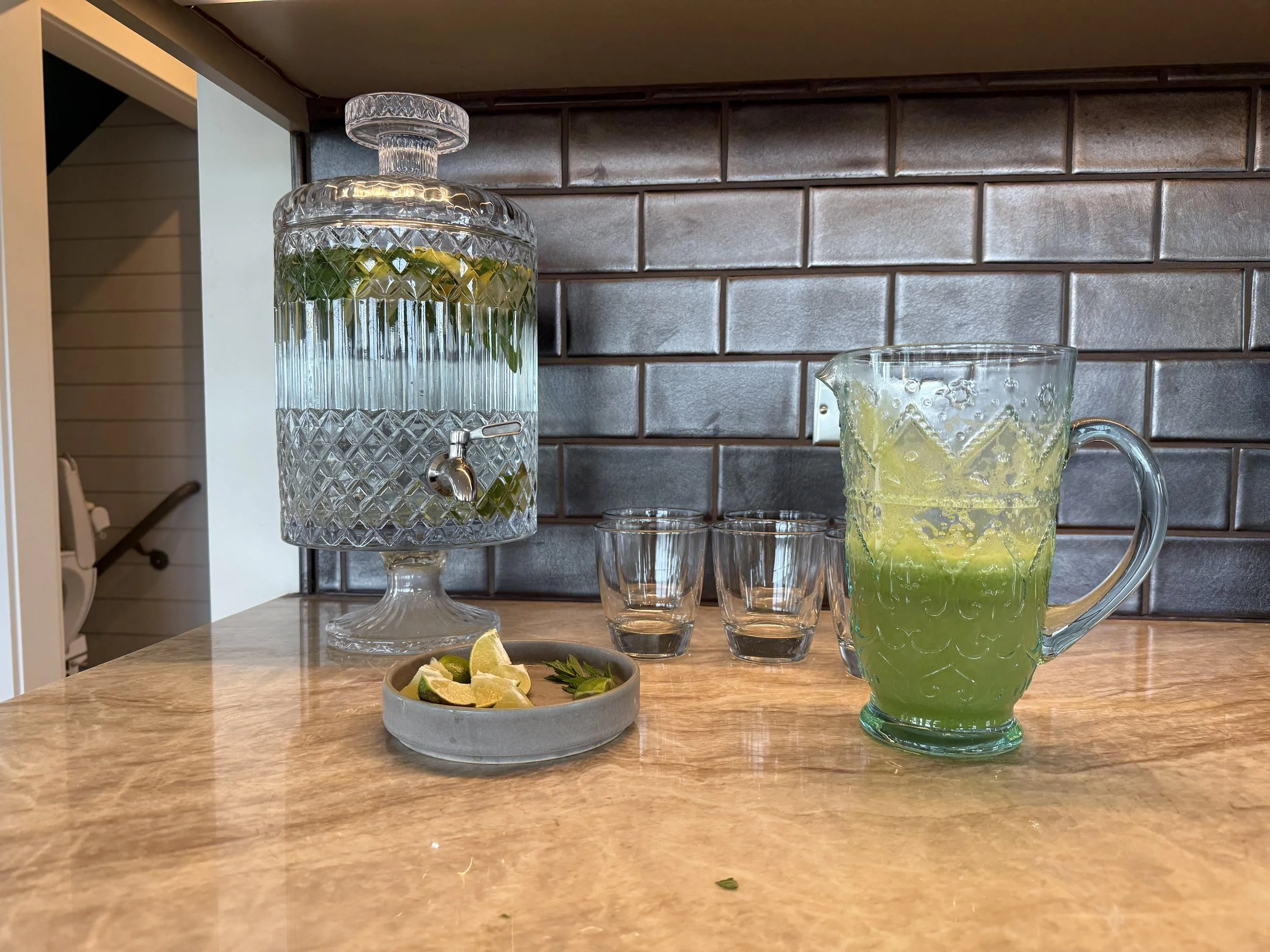 Glass beverage dispenser filled with water, lemon slices, and mint leaves; glass pitcher with lemon and lime slices; two small empty glasses; lemon and lime wedges on a small dish; tiled backsplash and wooden countertop.