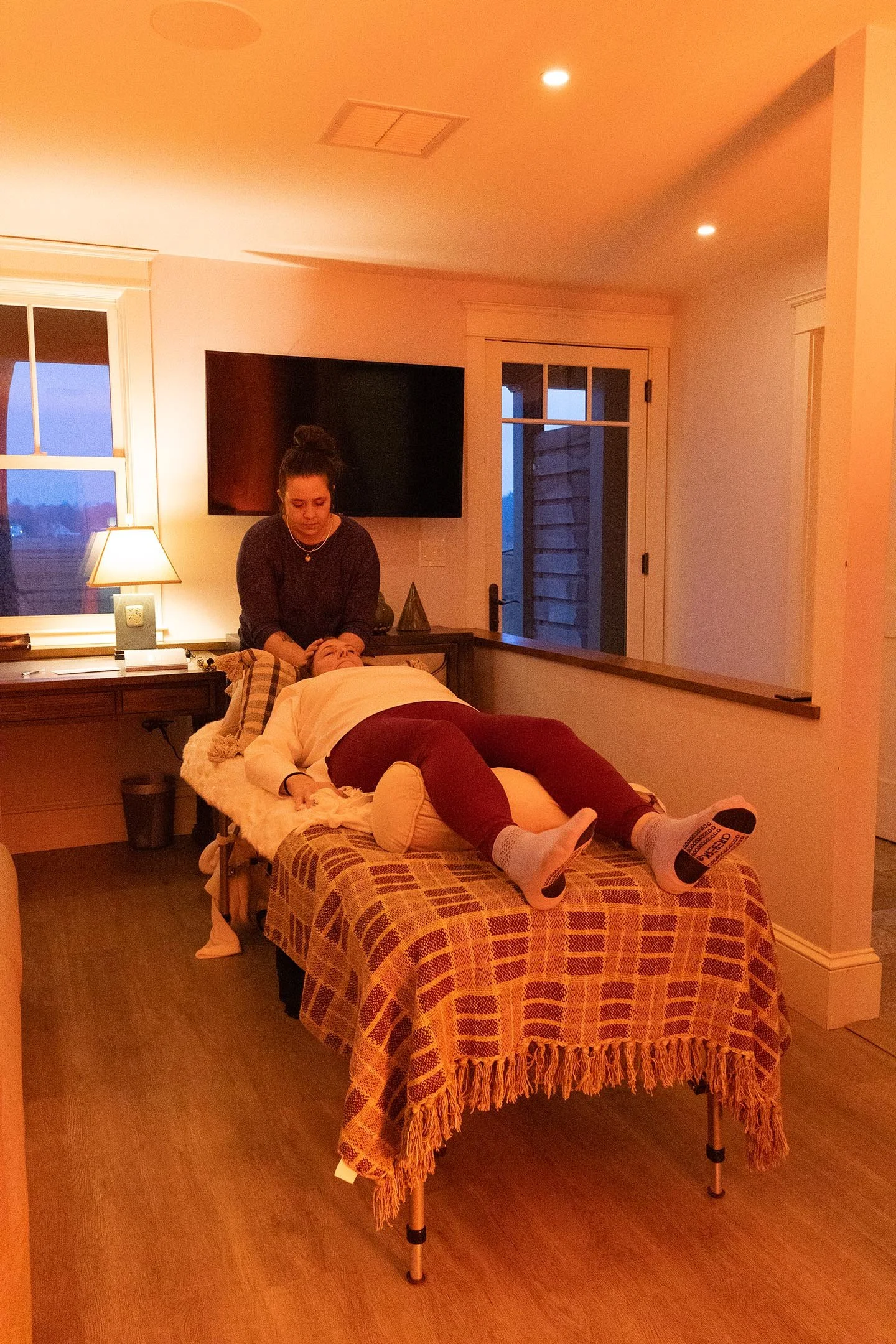 A wellness practitioner providing Reiki to a woman lying on a massage table in a cozy indoor setting with soft lighting, a window, and a door.