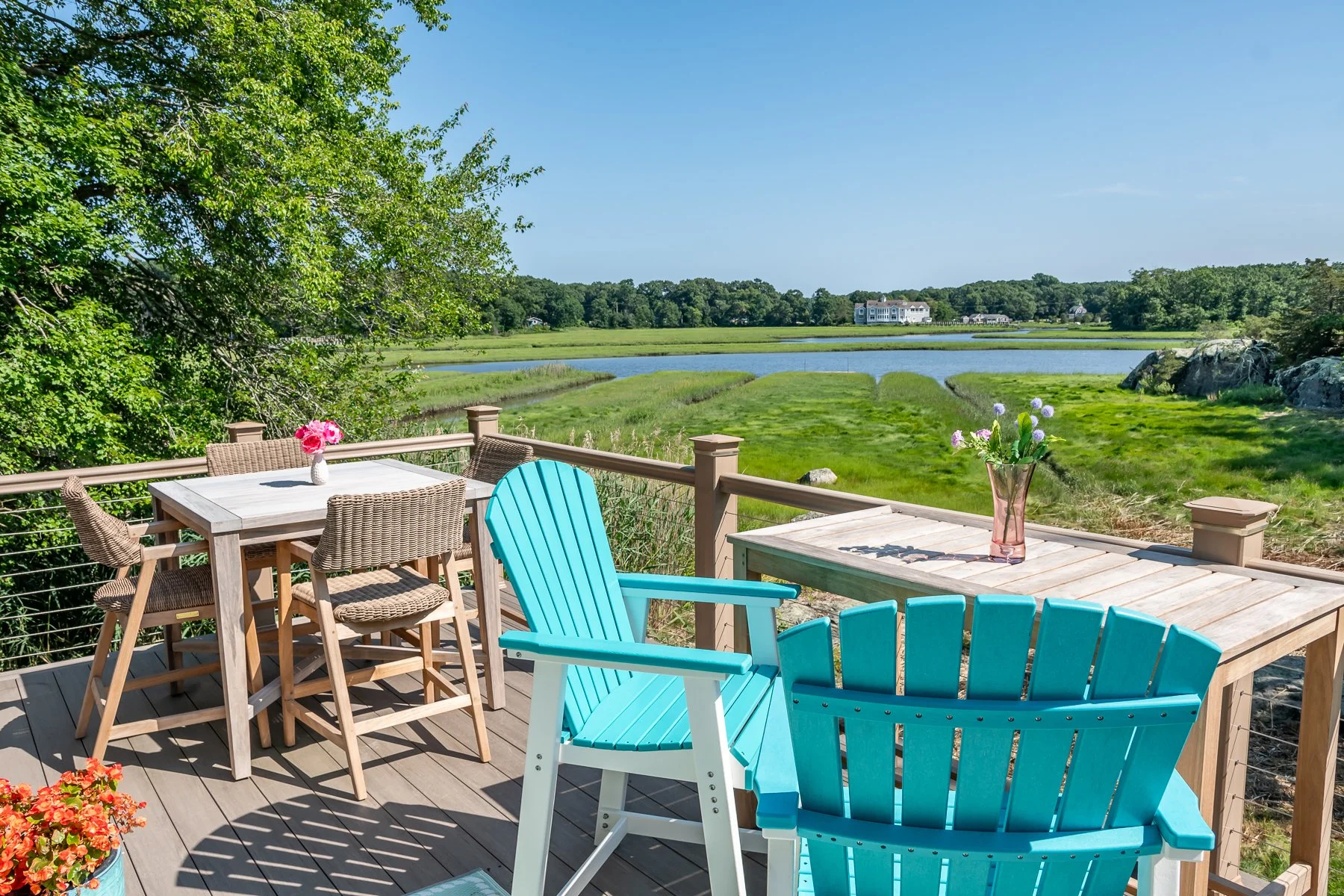 A deck overlooking a lush green landscape with a river, trees, and a house in the distance. There are outdoor furniture and a flower vase on the deck.