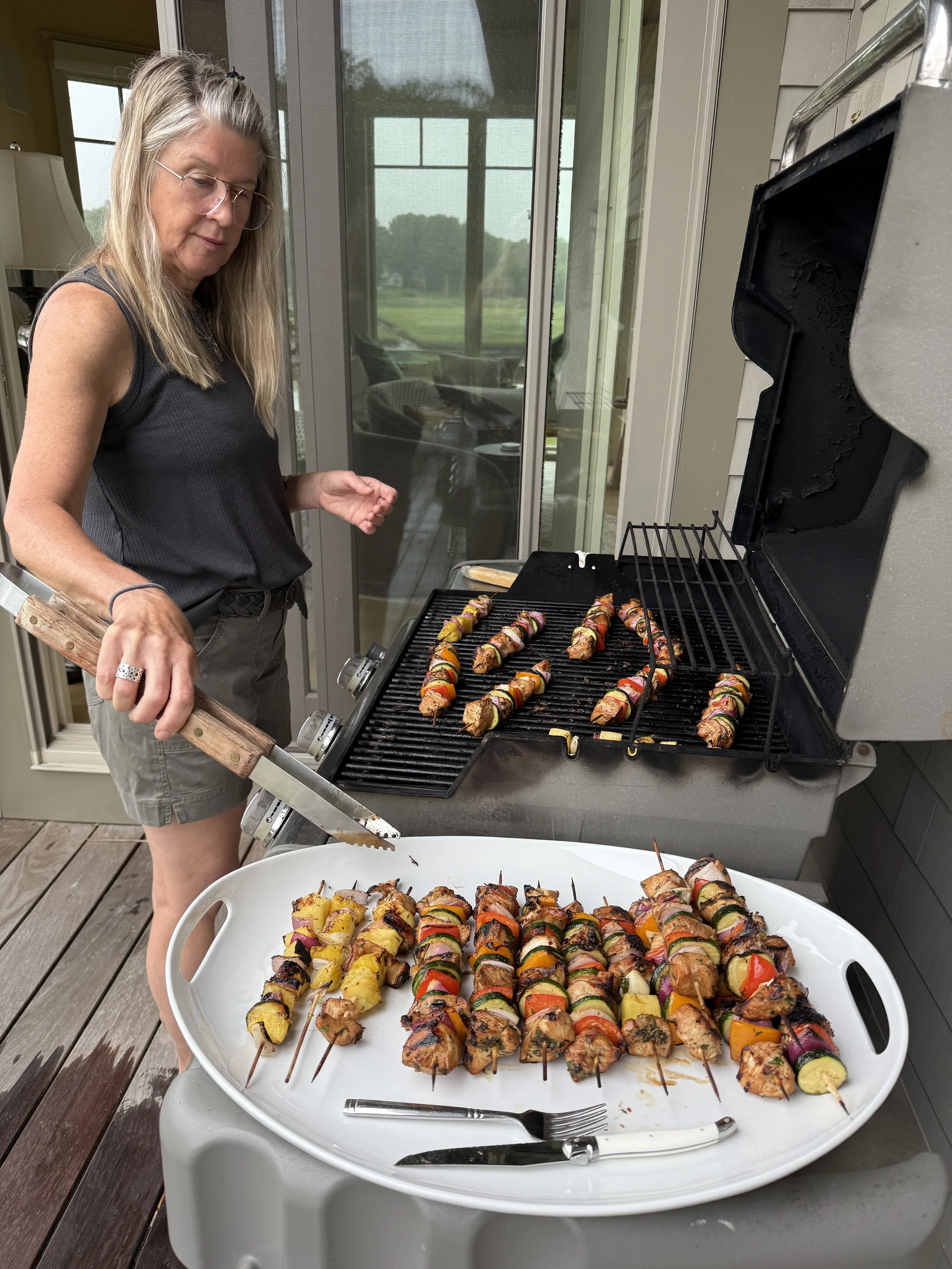 A woman grilling colorful vegetable and meat skewers on a barbecue grill outside on a deck.