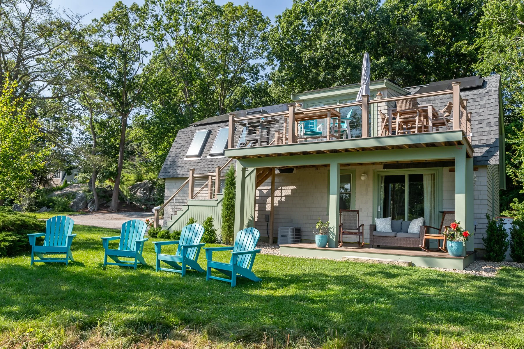 A two-story house with a backyard patio and a grassy lawn. The patio has outdoor furniture including a sofa and chairs, with a second story balcony above. Several turquoise Adirondack chairs are arranged on the lawn facing the house. Tall trees surro