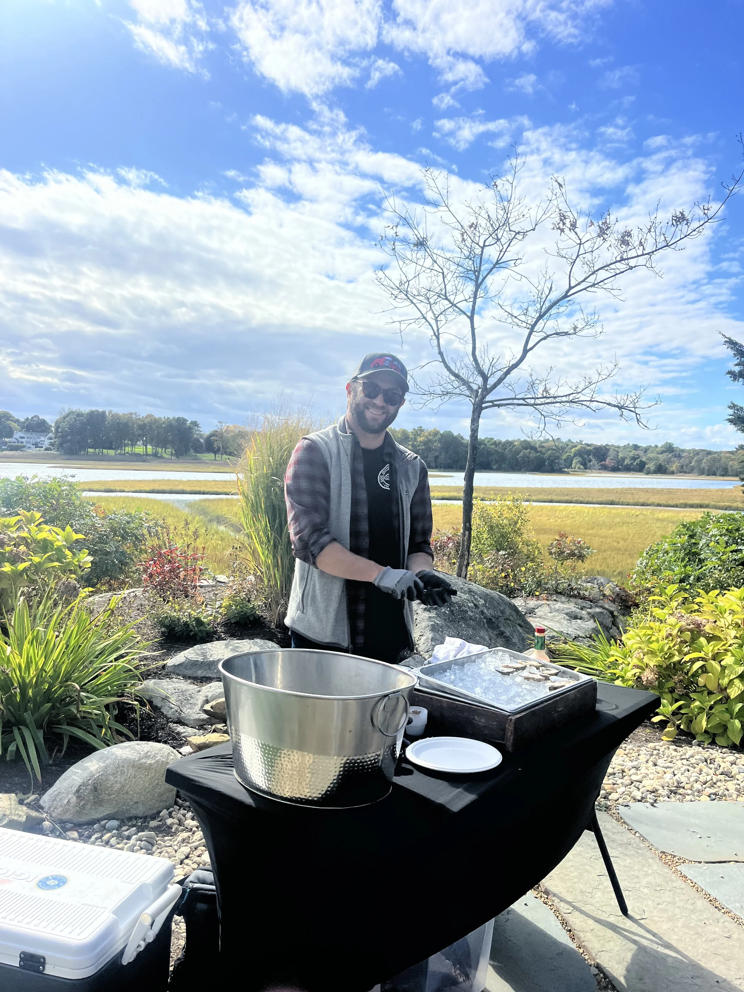 A man is grilling outdoors near a lake with a landscape of trees and a partly cloudy sky. He is smiling, wearing sunglasses, a black cap, a gray vest over a plaid shirt, and gloves, standing behind a black table with a grill and some supplies.