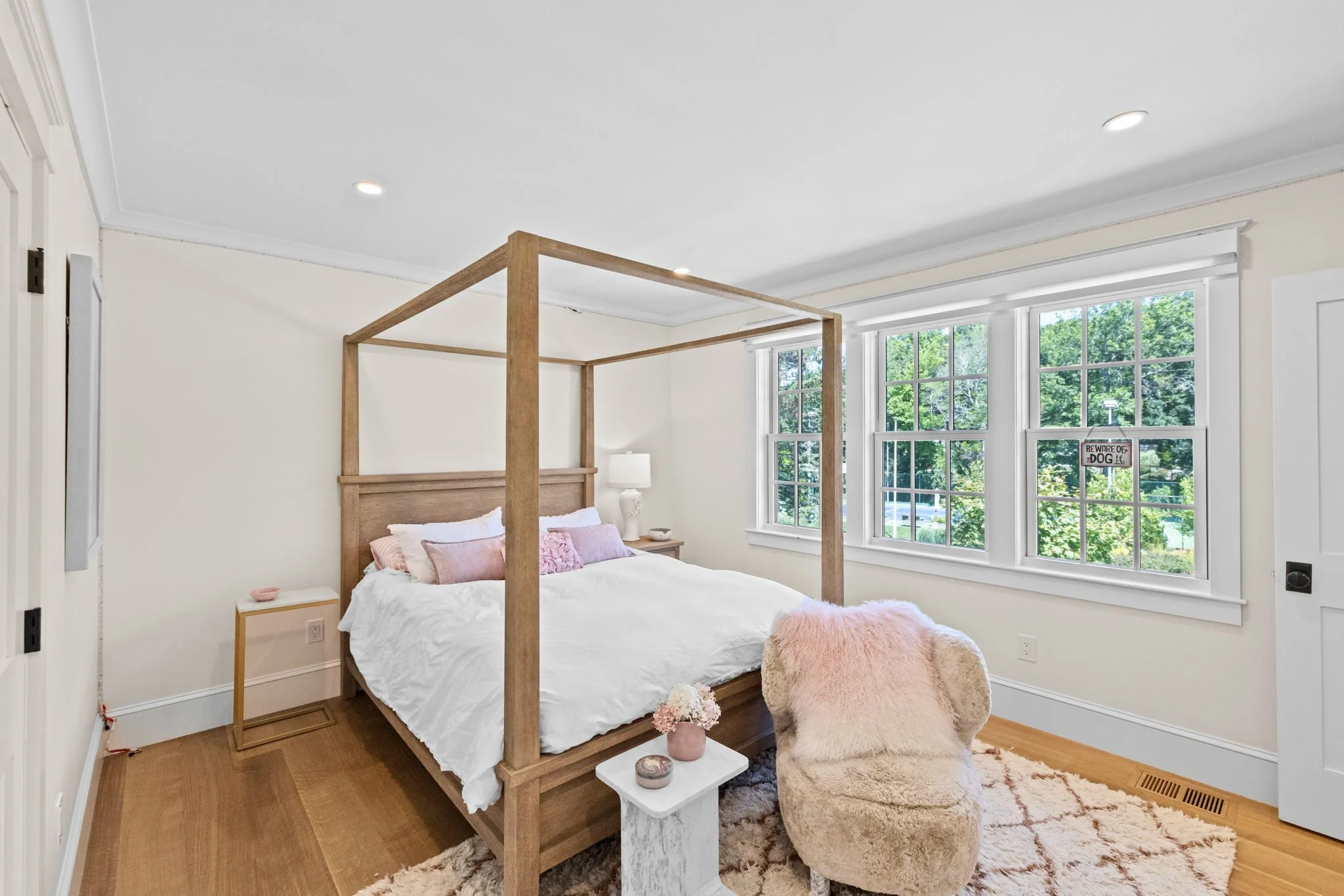Bright bedroom with a wooden four-poster bed, soft pink pillows,  a cozy chair, and large windows with a view of trees outside.