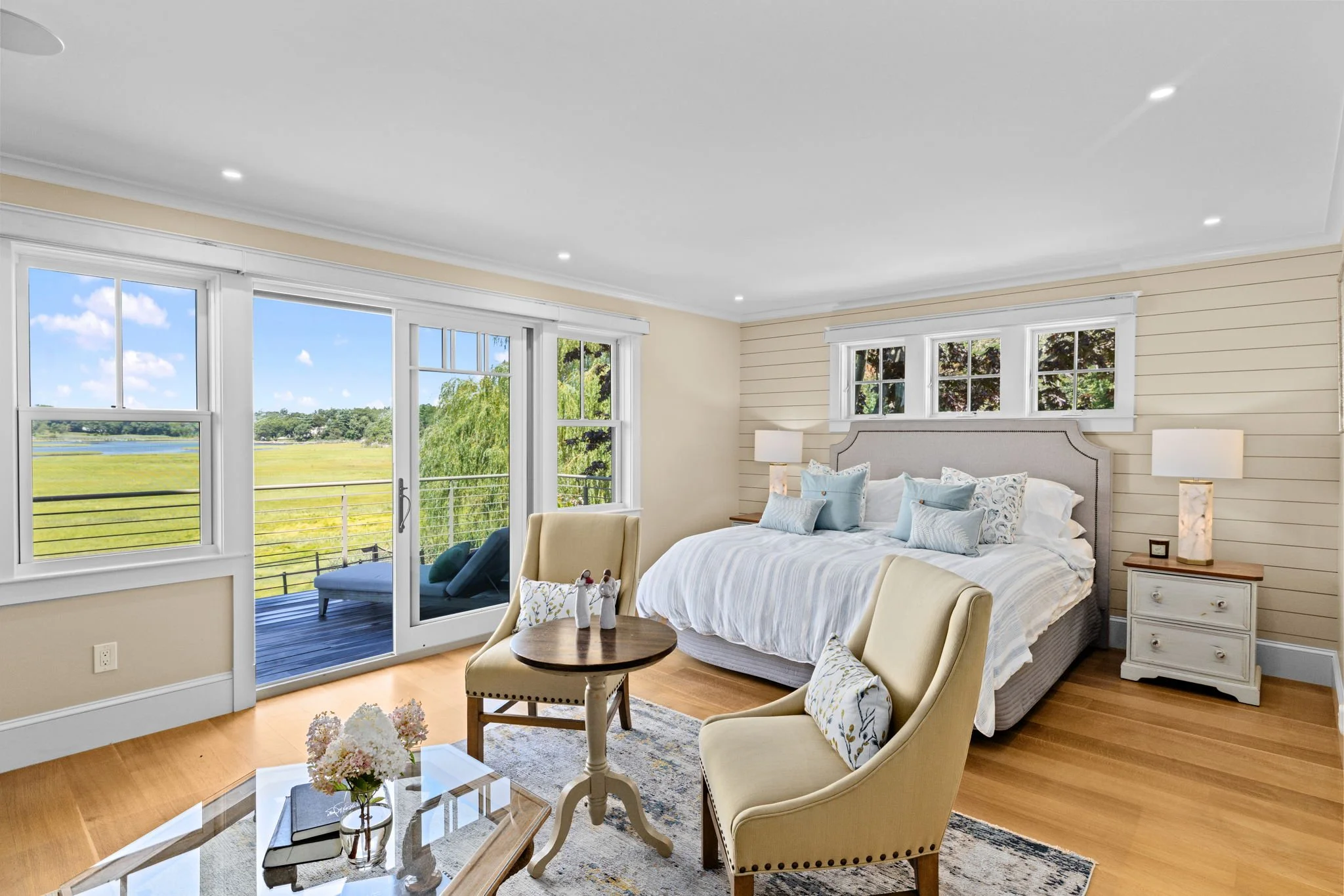 Bright bedroom with large windows view of outdoors, king-sized bed with white bedding and blue pillows, two cream armchairs with decorative pillows, small round wooden table, and bedside lamps, hardwood floor, and a sliding glass door leading to a balcony.