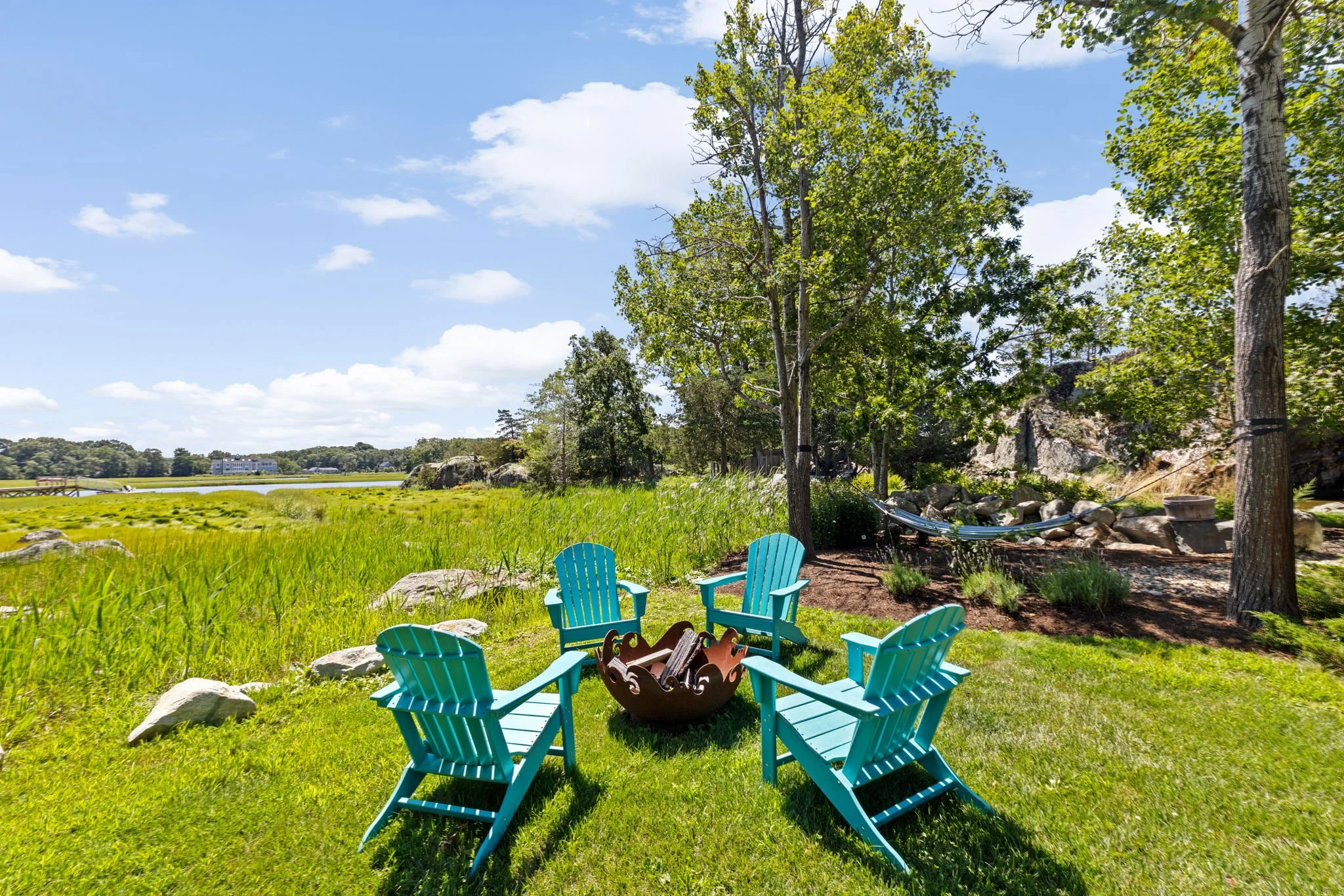 Four teal Adirondack chairs arranged in a circle around a fire pit on lush green grass in a scenic outdoor setting with trees, rocks, a hammock, and a body of water under a partly cloudy sky.