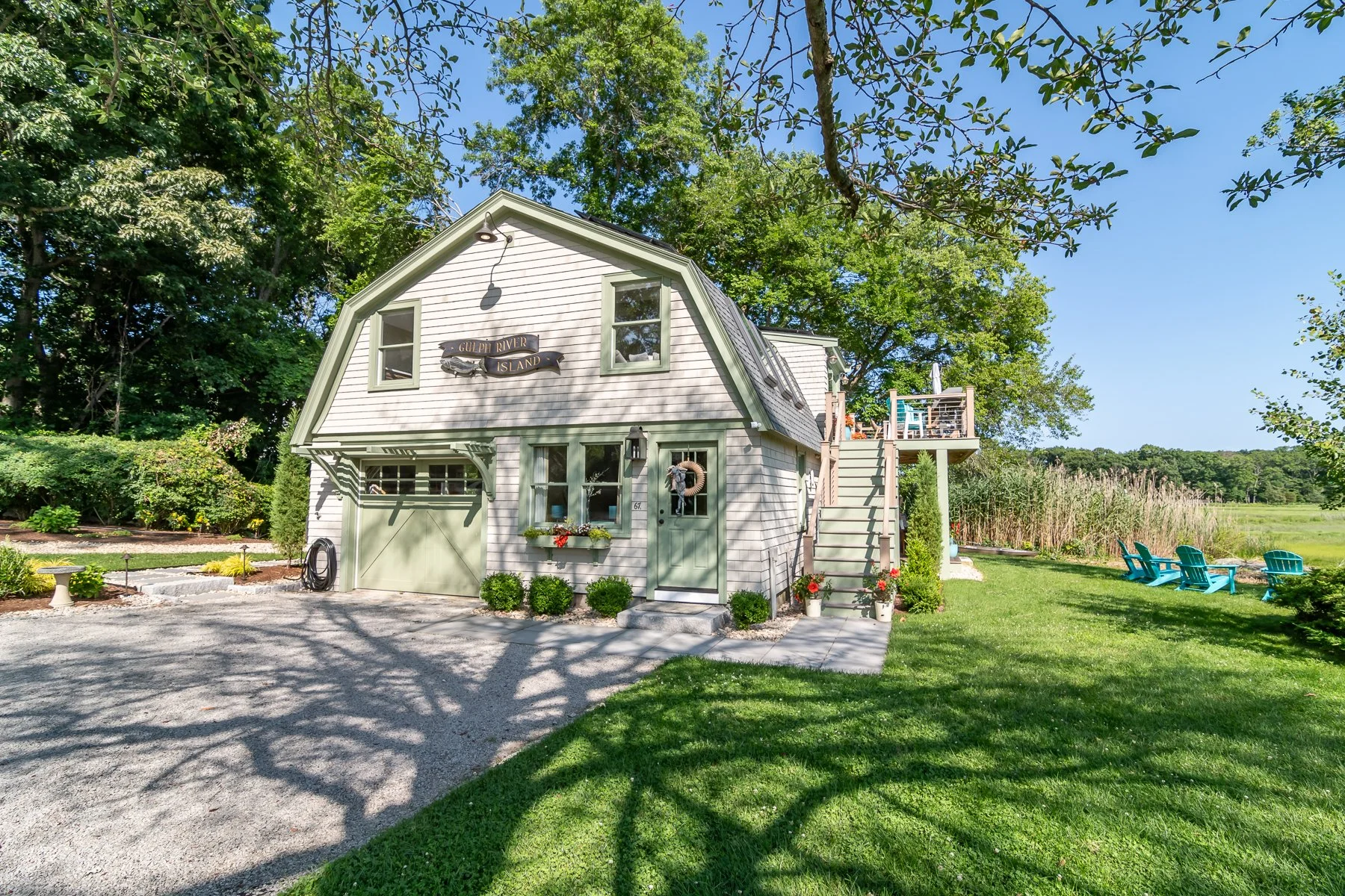 A charming two-story house with white and green exterior, surrounded by lush greenery, with outdoor seating and a gravel driveway, under a bright blue sky.