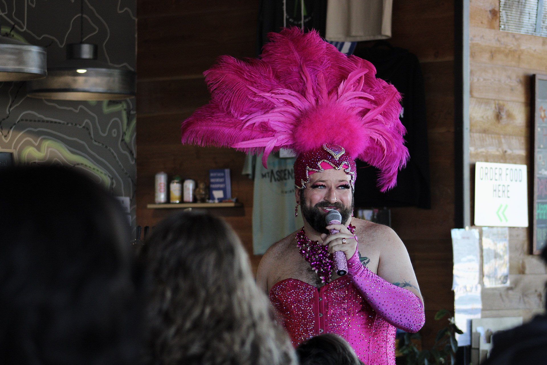 Avery DeLite dressed in a pink rhinestoned costume with a large pink feathered headdress, speaking into a microphone at an indoor burlesque show in Helena, Montana, with an audience in the foreground as a fundraiser for Montana Pride.