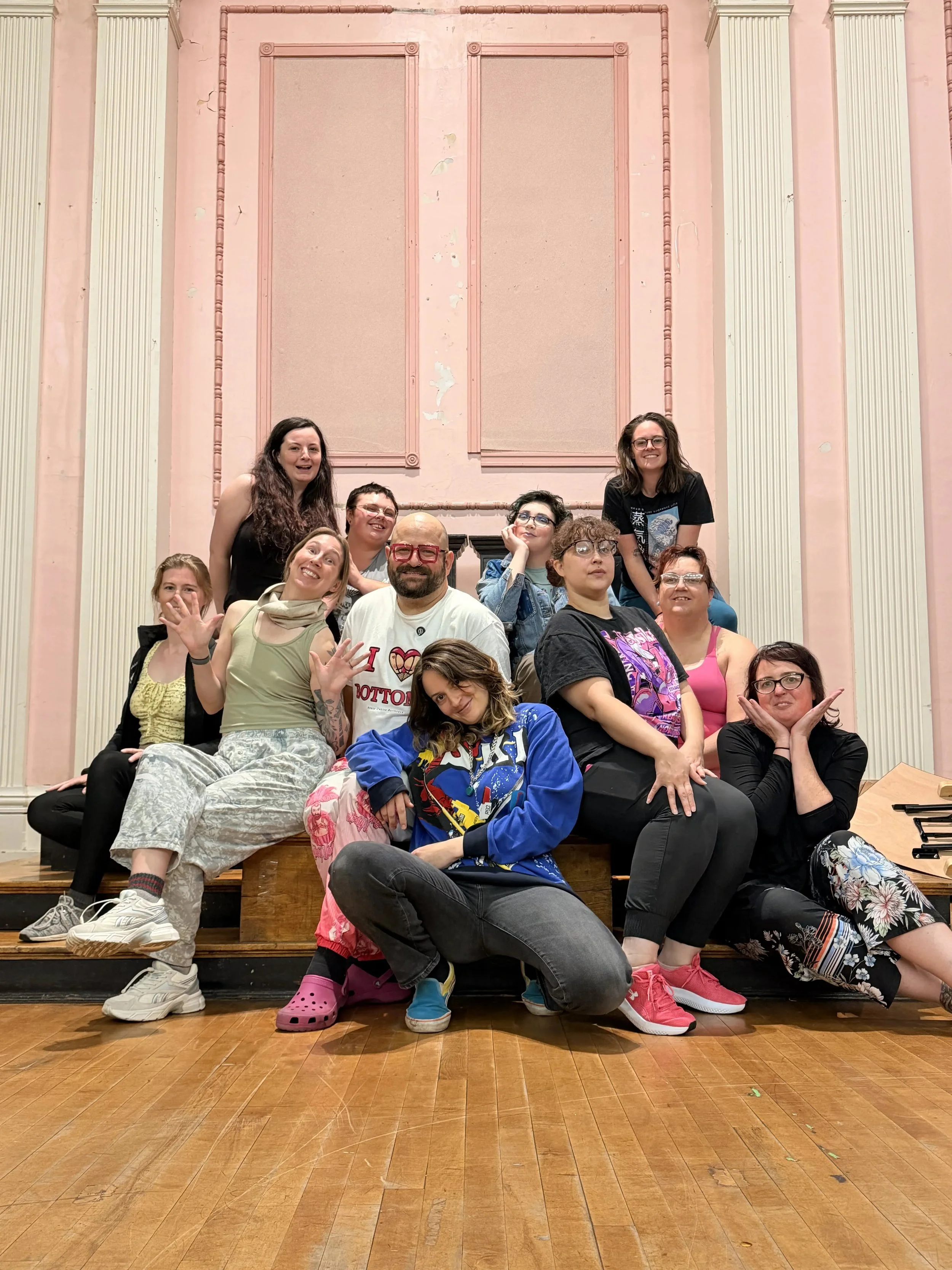 A group of people posing for a photo on stage in a theater or auditorium, with a pink wall and ornate trim in the background. They are smiling and making playful gestures, some sitting and some standing.