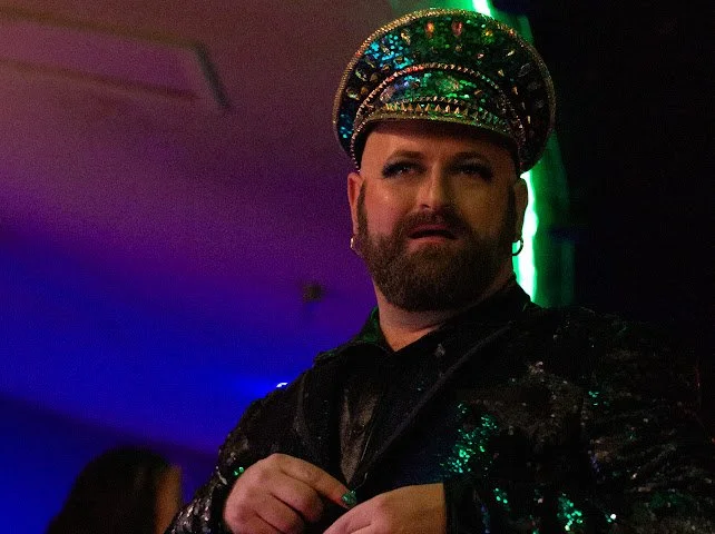 Avery DeLite wearing a colorful, sparkly hat and a green sequined jacket, performing in front of a dark backdrop with neon lighting during a Missoula, Montana burlesque show. Photo by Isa Tumble.