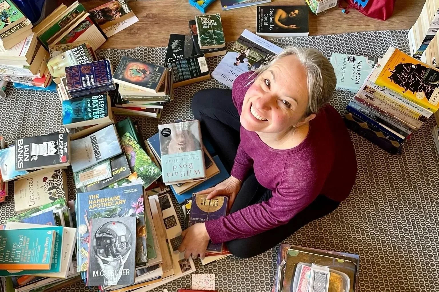 Professional York home organiser Cher Casey smiling and wearing a maroon top, sitting on the living room floor surrounded by books to declutter and organise.