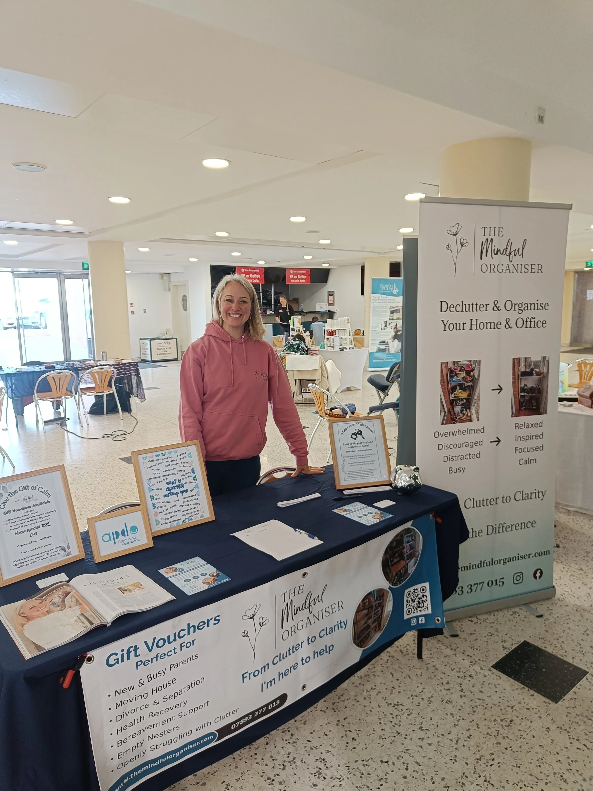 Cher Casey, York's decluttering expert, in a pink hoodie standing behind a table with informational pamphlets and a banner for The Mindful Organiser, offering decluttering and organisation services for home and office, in York's Racecourse building