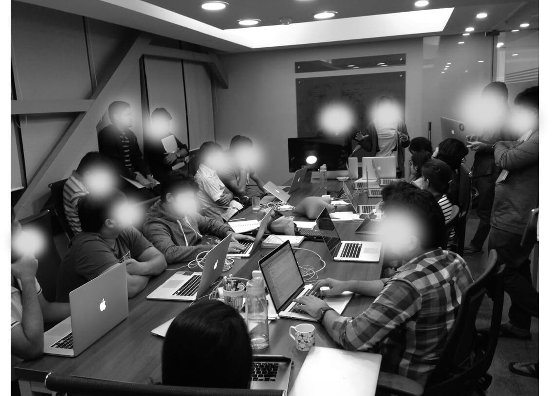 A group of people in a meeting room working on laptops around a large conference table. Several individuals are standing, some presenting or discussing with others seated. The room has a modern design with overhead lighting.
