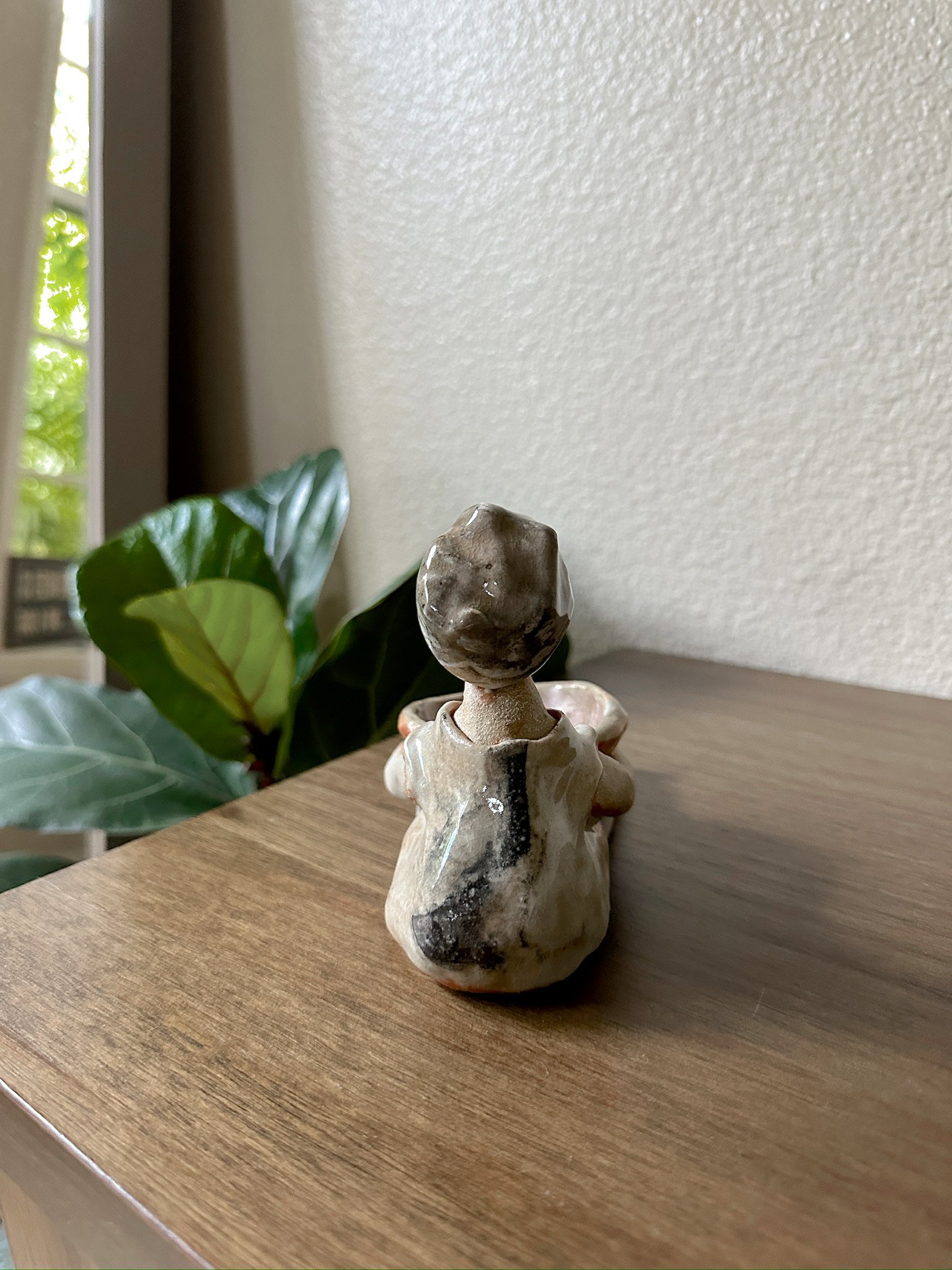 Glazed and Fired ceramic handmade sculpture of a lady holding a light red bowl and sitting on a table. Image from the back.